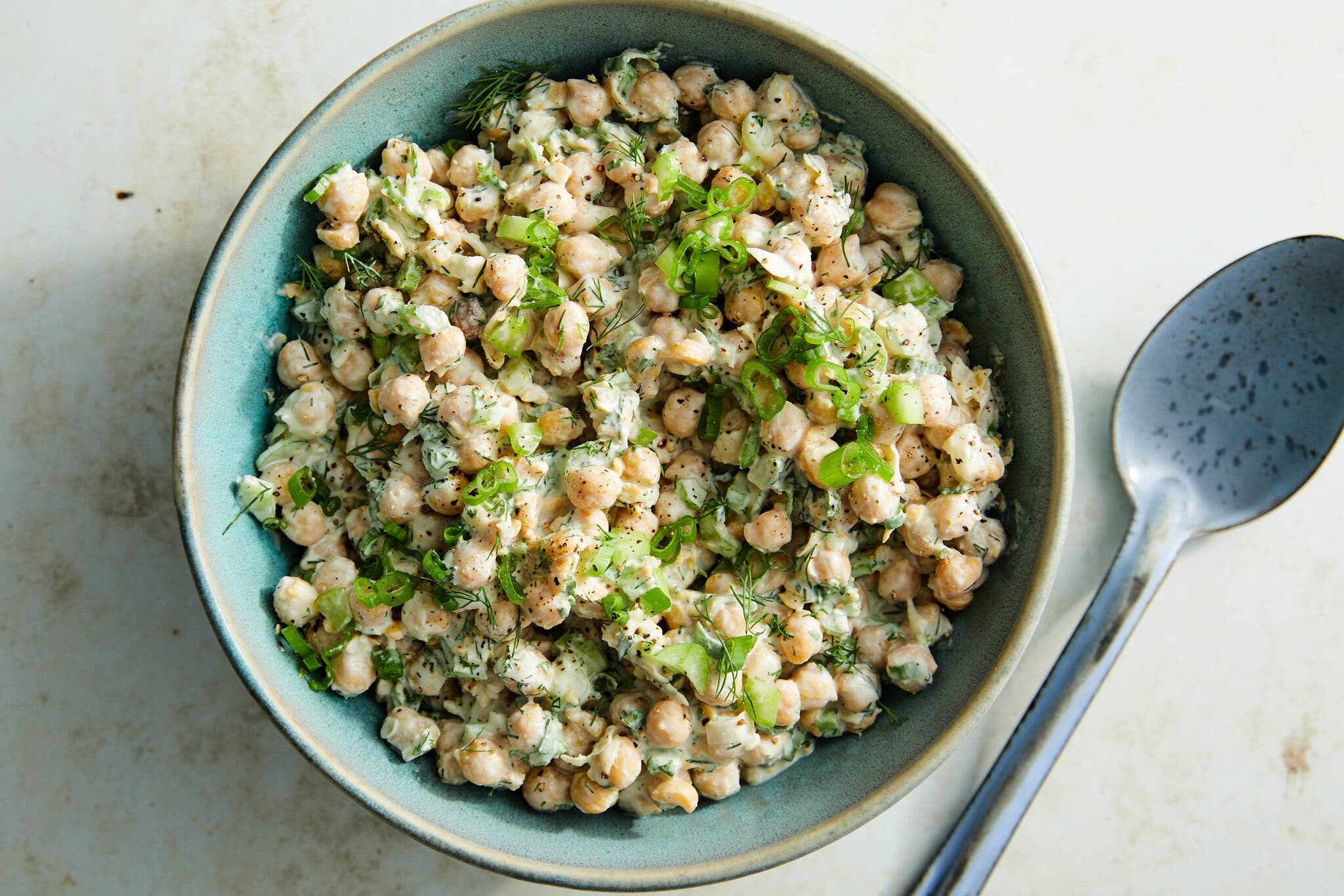 Chickpea salad topped with dill, parsley and scallions in a light blue bowl. To the right is a serving spoon. 