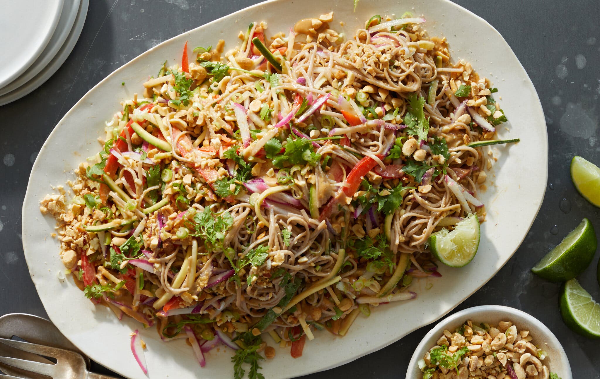 A white dish holds soba salad with spicy peanut sauce, topped with chopped peanuts and fresh herbs. To the right are some lime wedges.