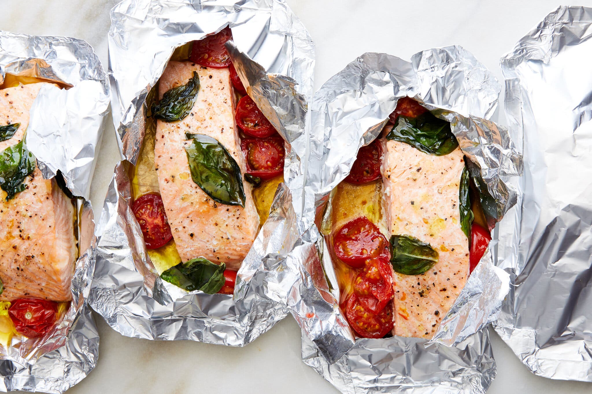 An overhead shot of three packets of salmon wrapped in aluminum foil, with tomatoes, basil and olive oil.