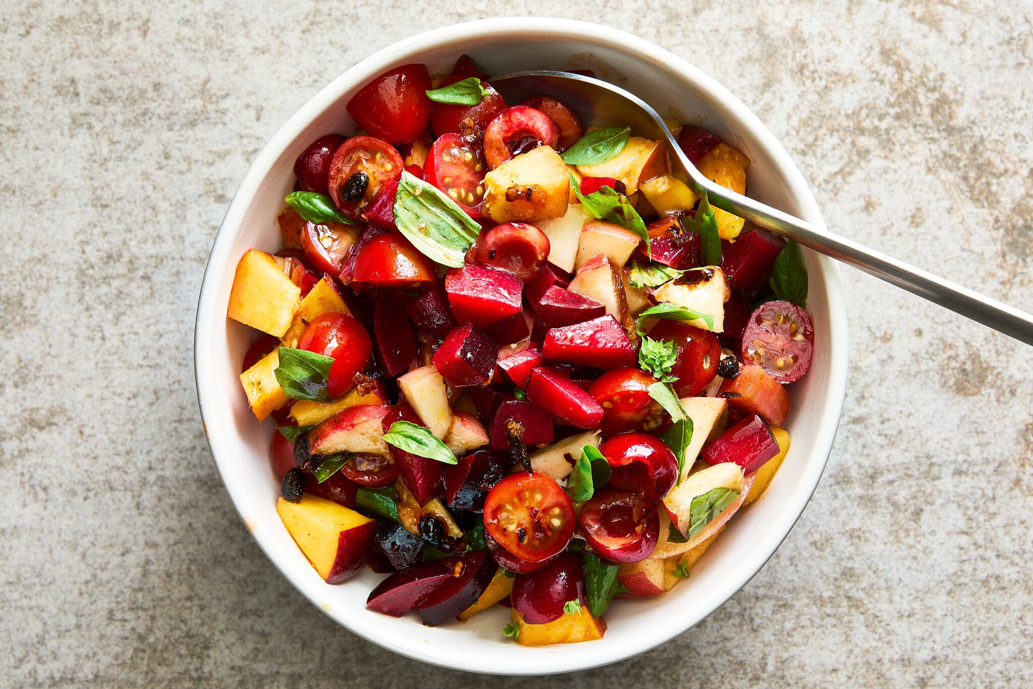 A bowl with cut stone fruit and cherry tomatoes topped with torn basil. 