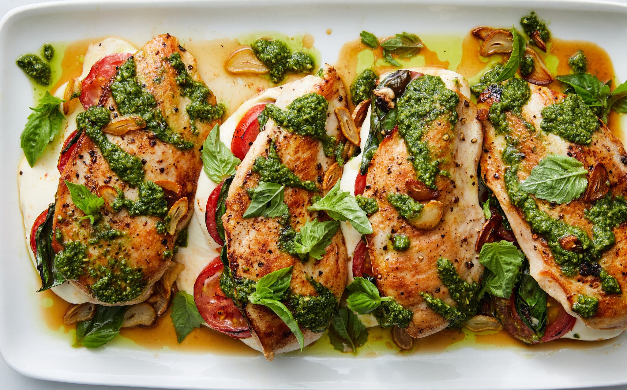 An overhead shot of a long rectangular serving plate with four large chicken breasts which are filled with sliced tomatoes, basil and melted mozzarella. They are garnished with pesto and browned on the top.