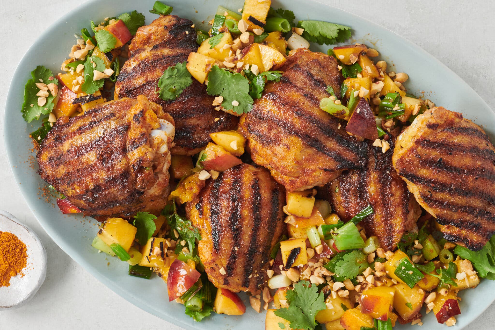 Six grilled suya spiced chicken thighs are nestled into a platter of grilled and chopped nectarines with herbs.