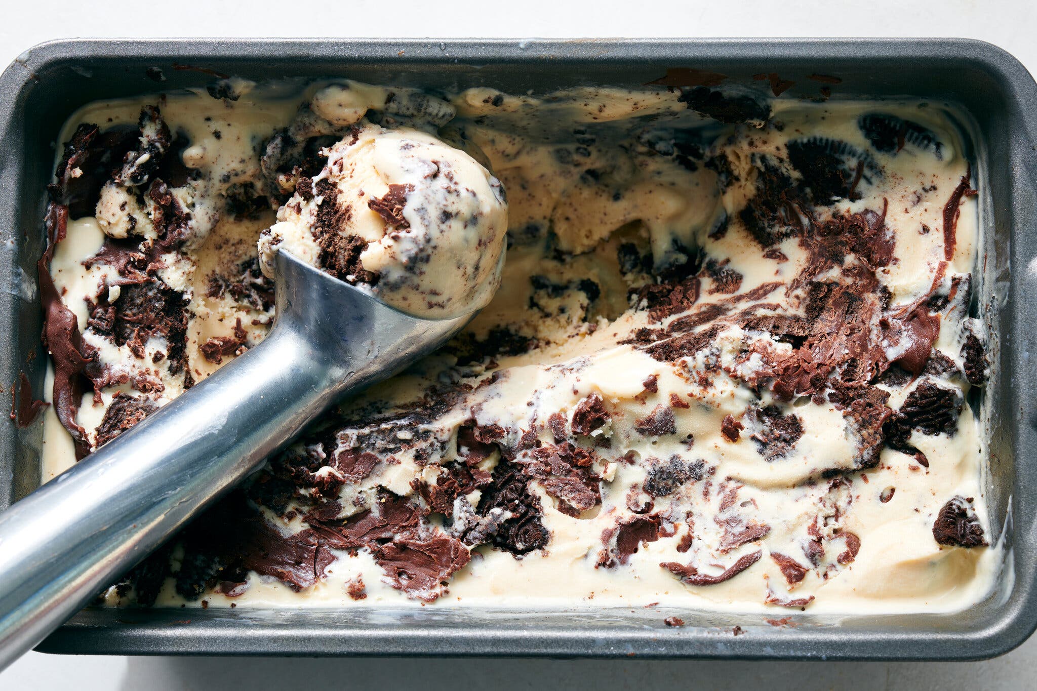 A metal ice cream scoop is filled with chocolate-swirled ice cream in a loaf pan.