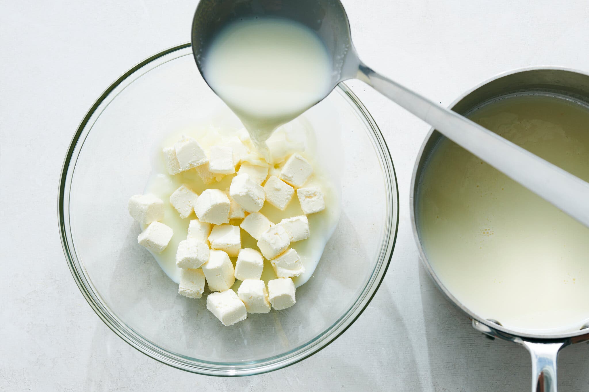 A ladle of milk from a small saucepan is being poured over cubes of cream cheese in a glass bowl.