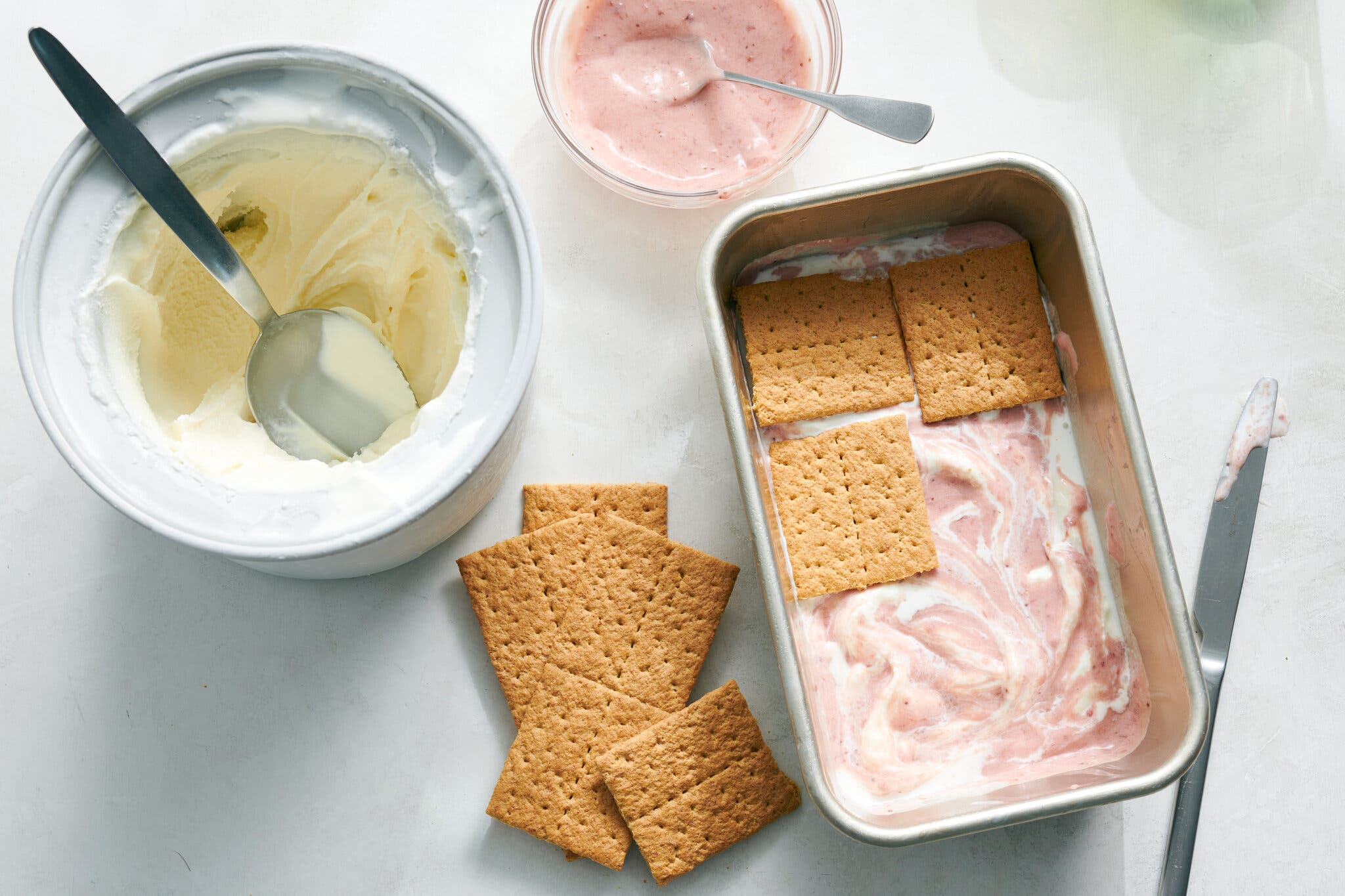 An ice cream maker filled with vanilla sits next to a bowl with strawberry jam and a metal loaf pan of the swirled ice cream with graham crackers on top.