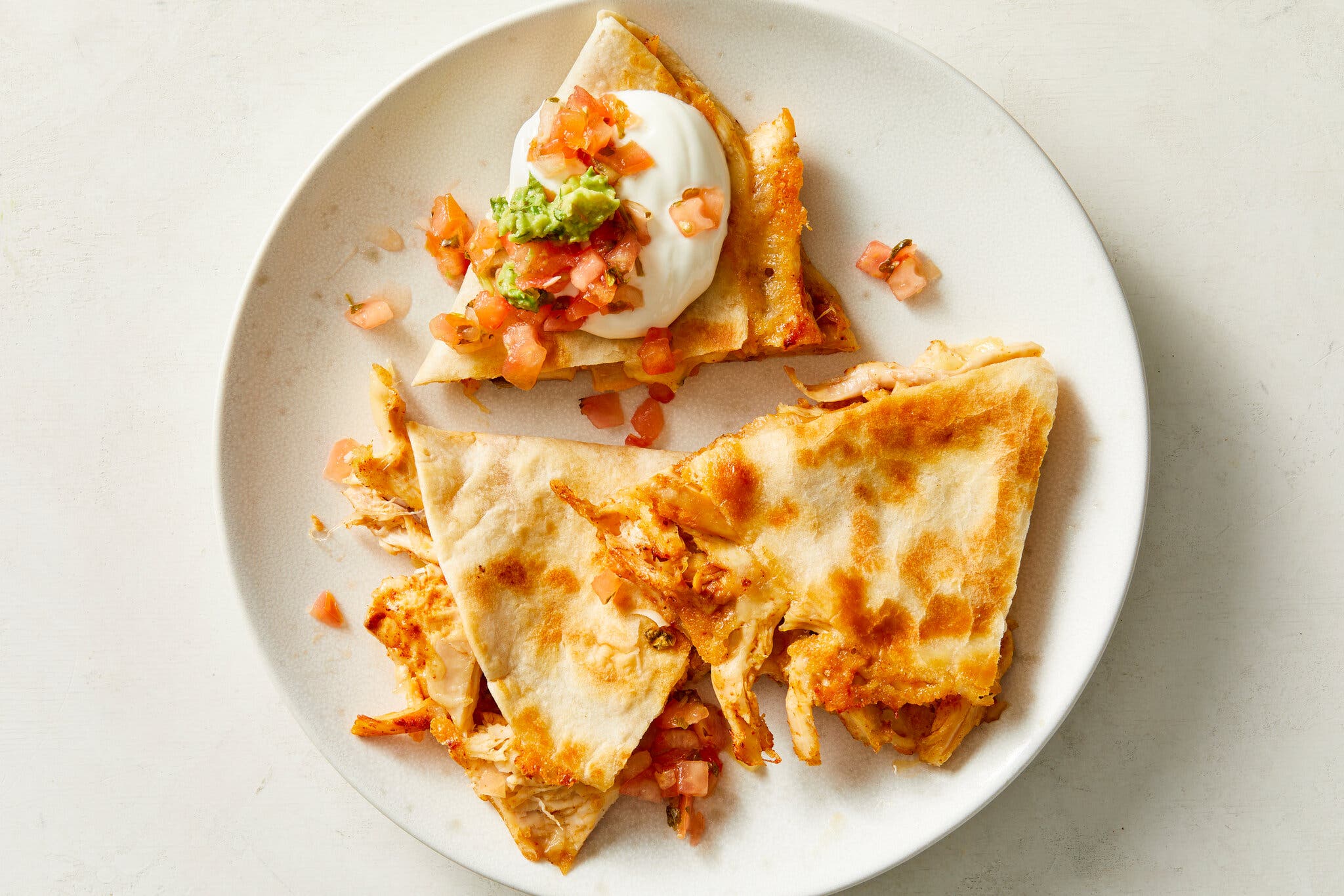 An overhead image of a white plate with three triangles of chicken quesadilla. One of them is topped with sour cream, pico de gallo and guacamole.