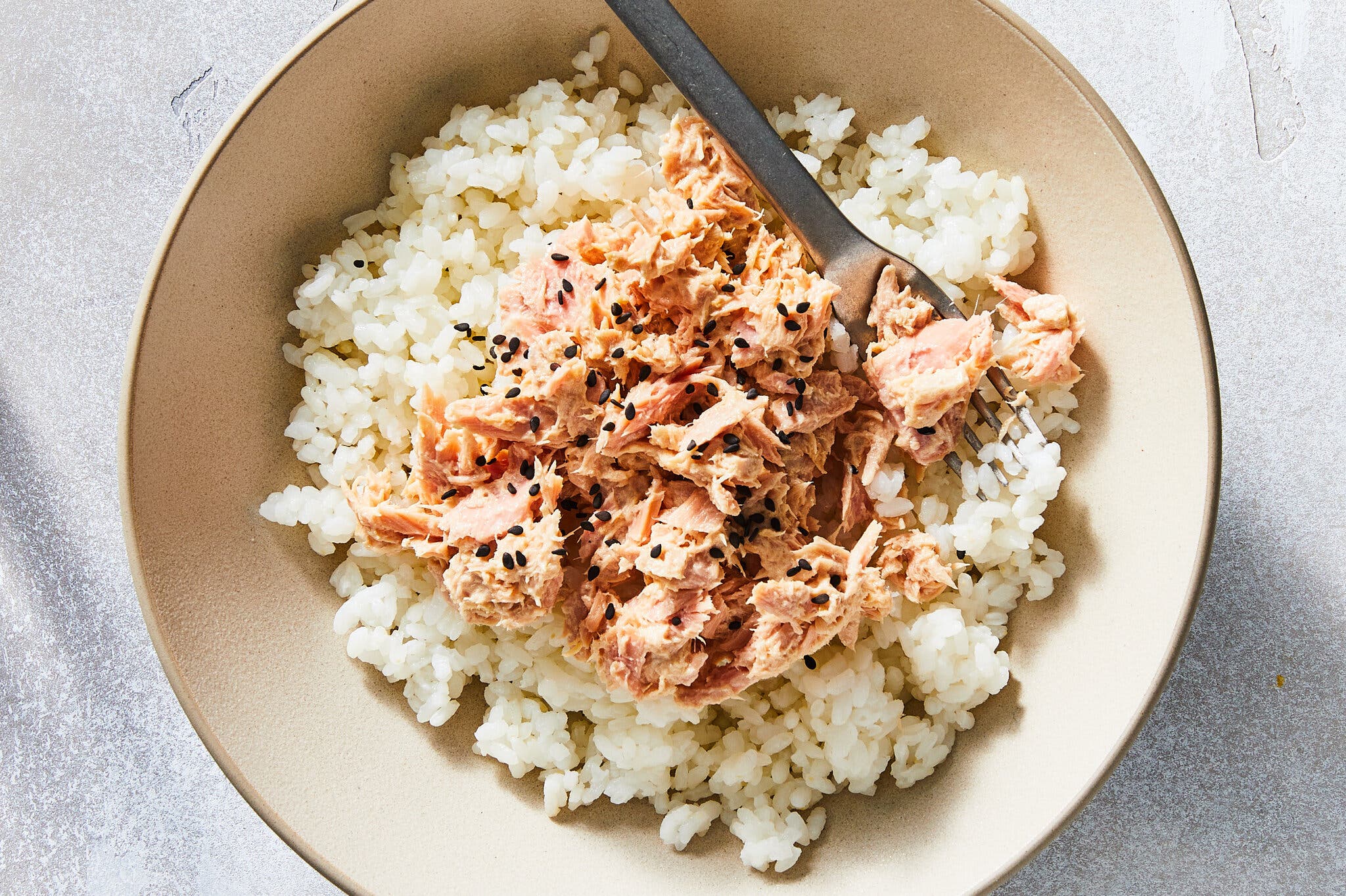 An overhead image of a bowl with white rice topped with canned tuna and black sesame seeds.