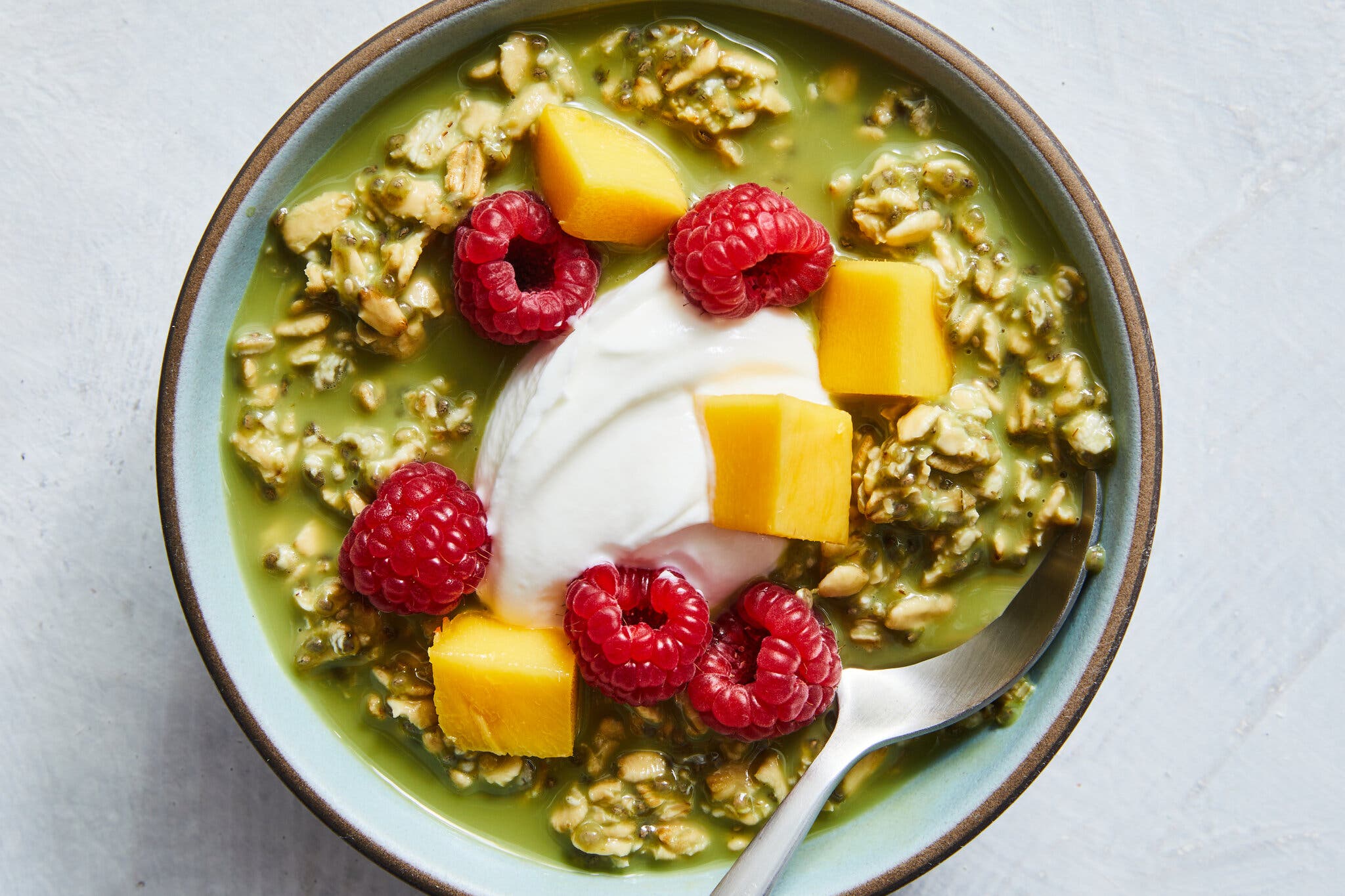 An overhead image of a bowl with bright green matcha overnight oats, topped with a dollop of yogurt, raspberries and mango.