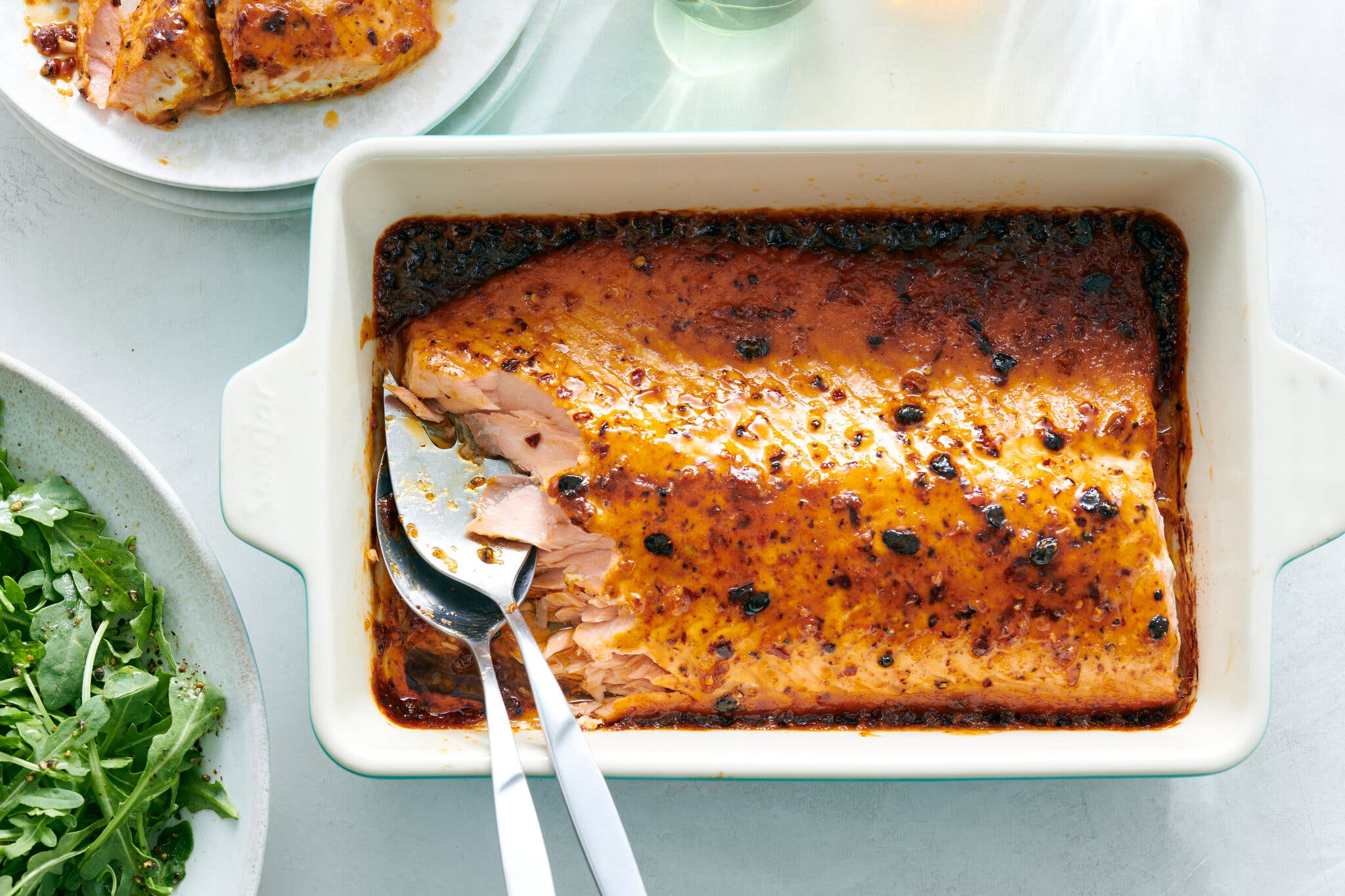 An overhead image of a baking tray with chile crisp and honey roasted salmon. At the bottom left corner of the frame sits an arugula salad.