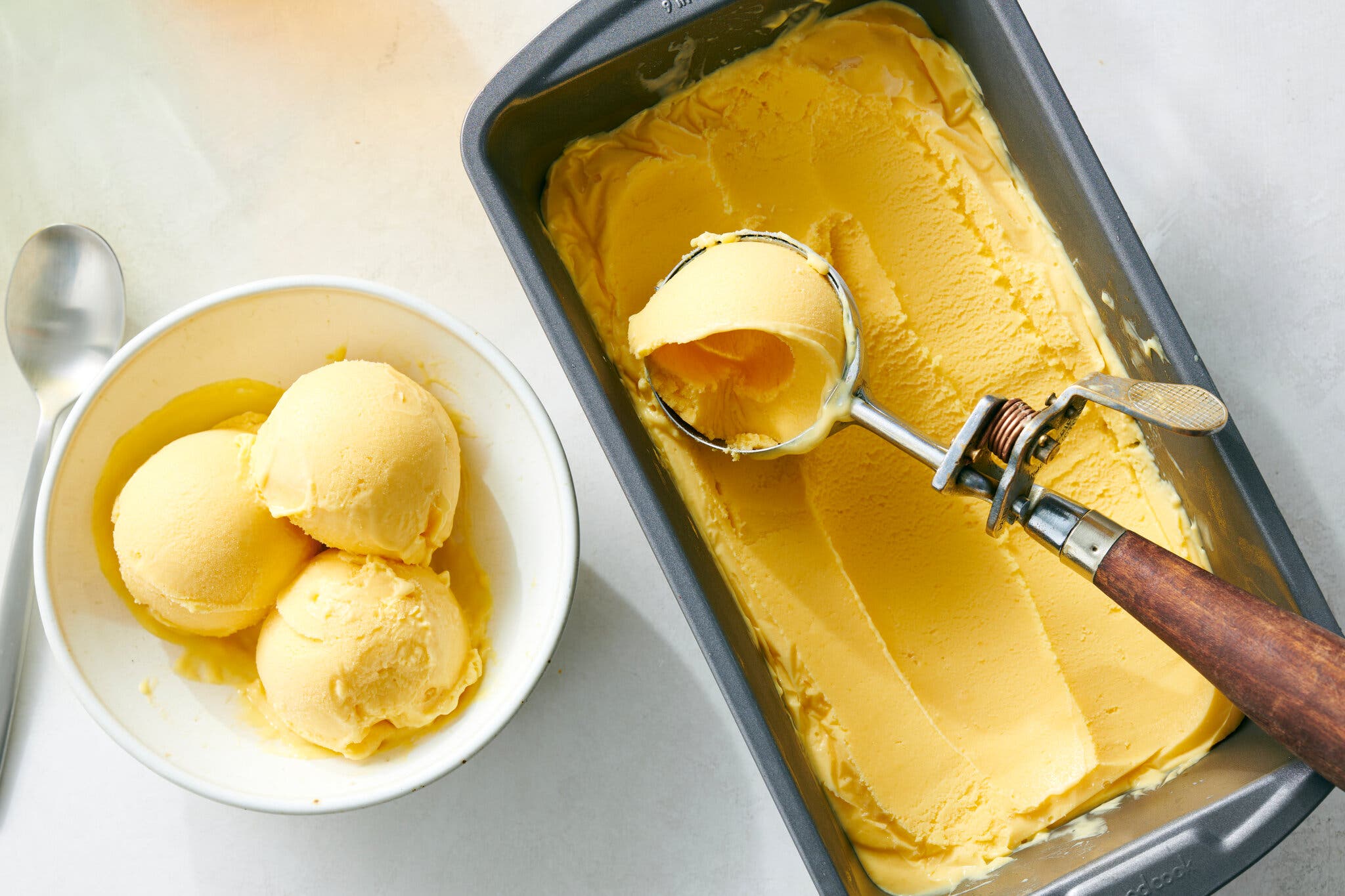 A loaf pan holds mango soft serve with an ice-cream scoop; three scoops of mango soft serve are next to it in a white bowl.