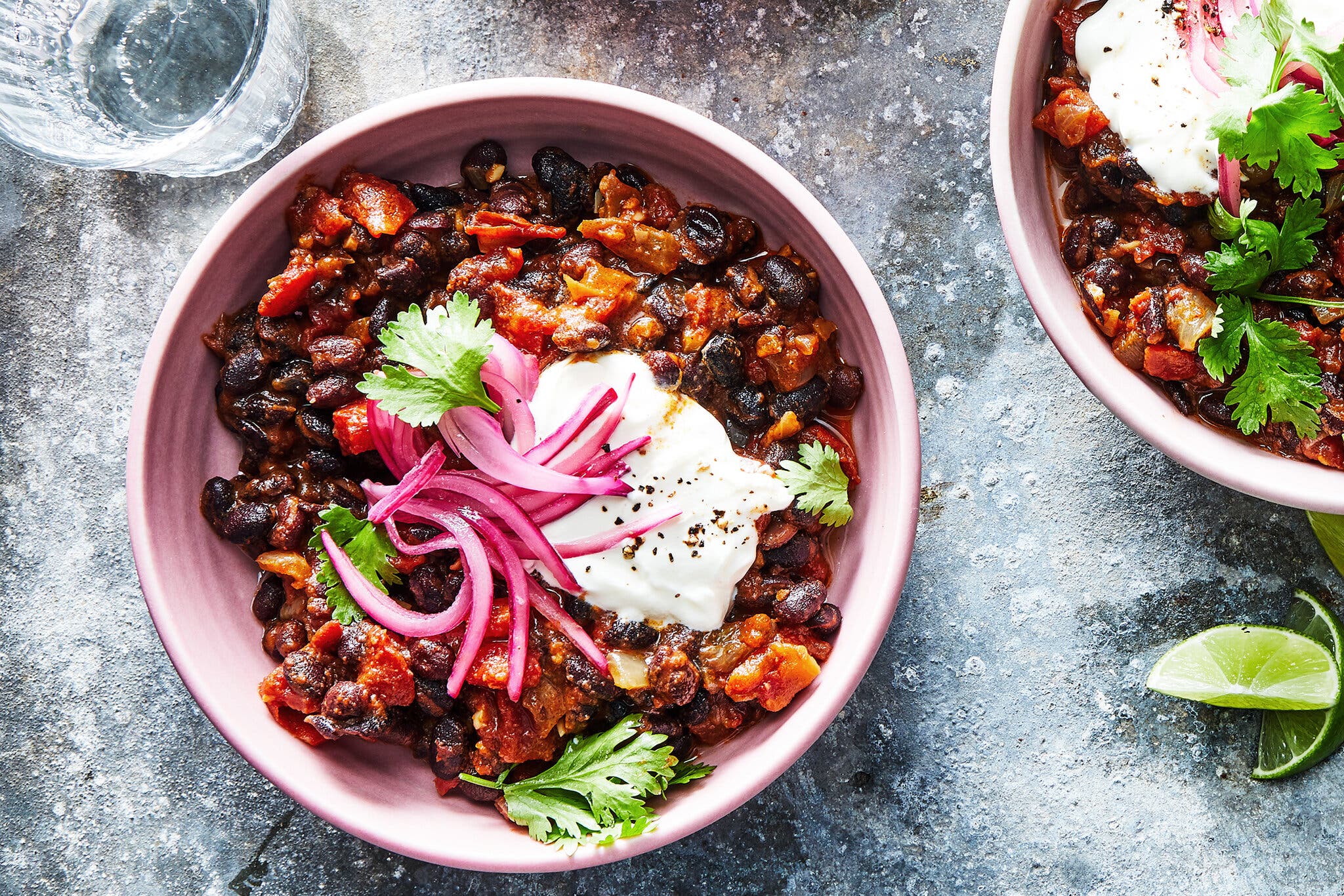 An overhead image of a pink bowl with vegetarian skillet chili topped with sour cream, pickled red onions and cilantro. To the right of the frame is another bowl of the same, cut off.