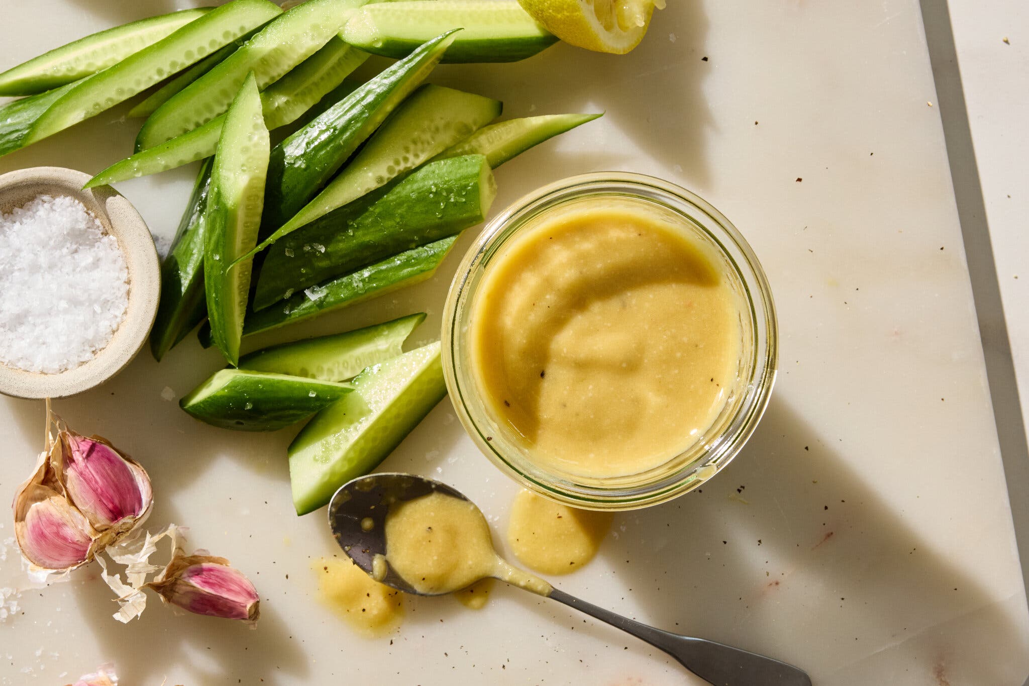 An overhead image of a cutting board with a bowl of olive oil next to cucumbers, salt and garlic.