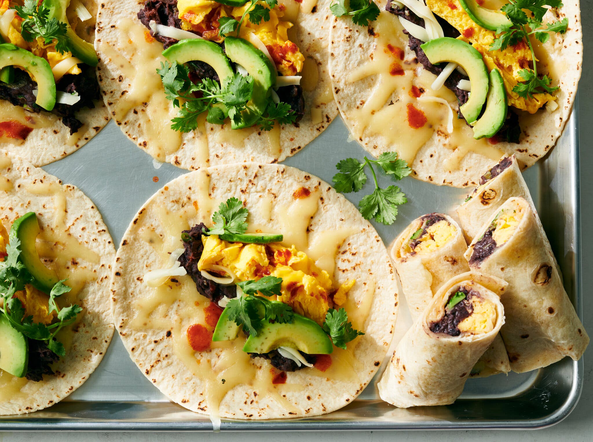 Six small tortillas topped with cheese, eggs, beans and avocado are photographed from overhead.