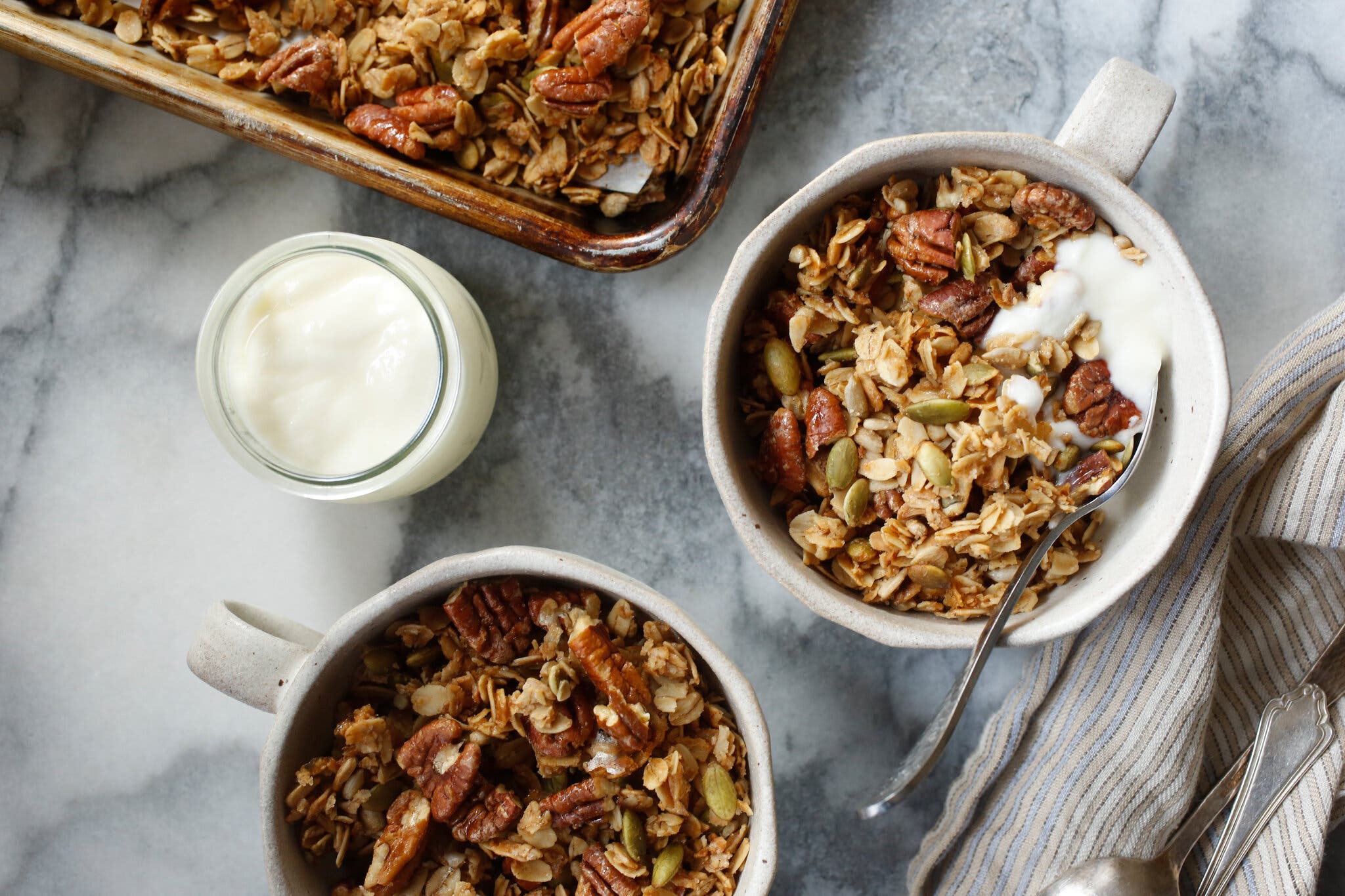 Two mugs are topped with granola. A small jar of yogurt sits to the side, as does a sheet pan filled with more granola.