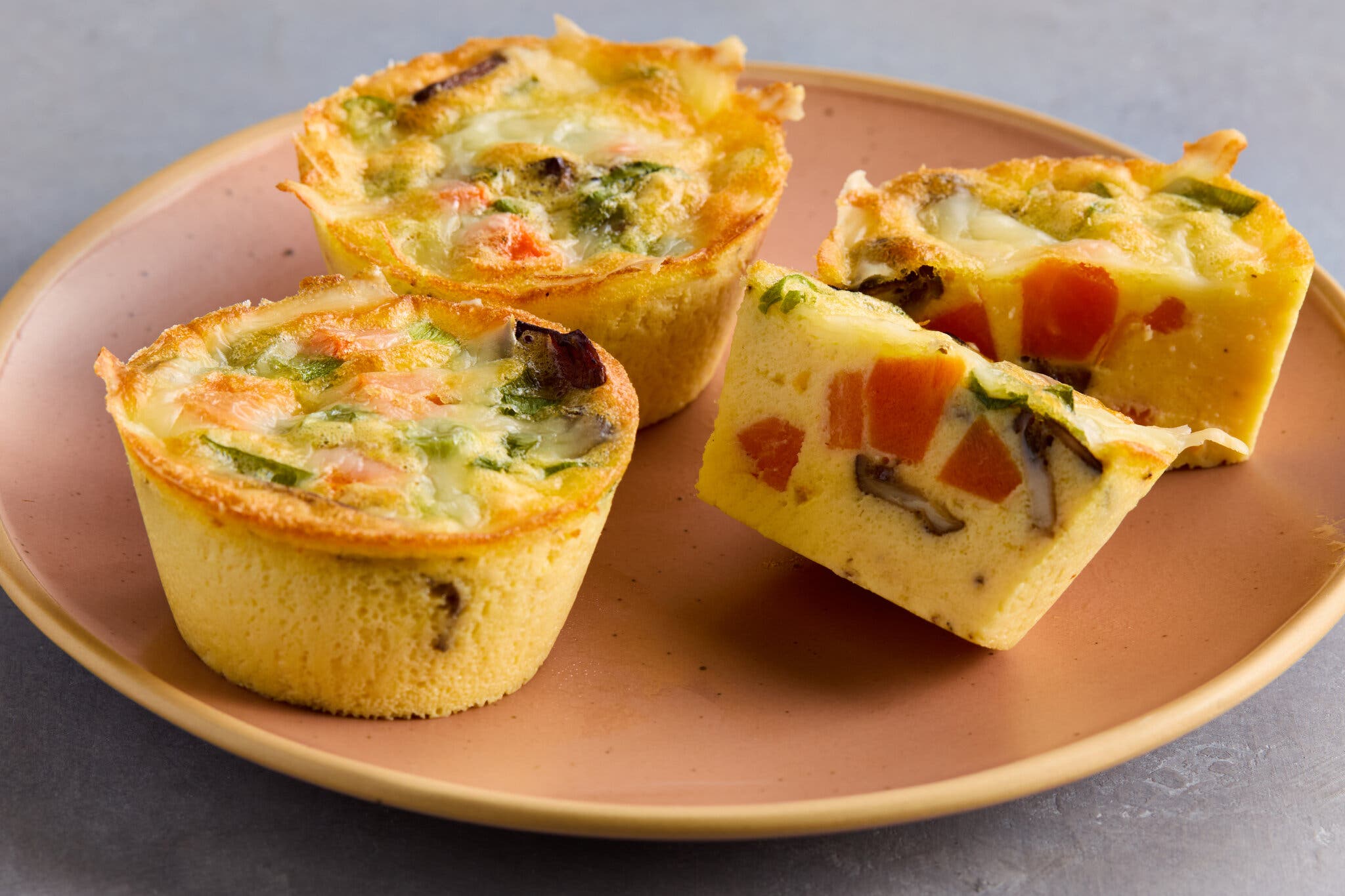 Three egg bites are photographed from the side on a plate. One is halved to show the vegetables inside.