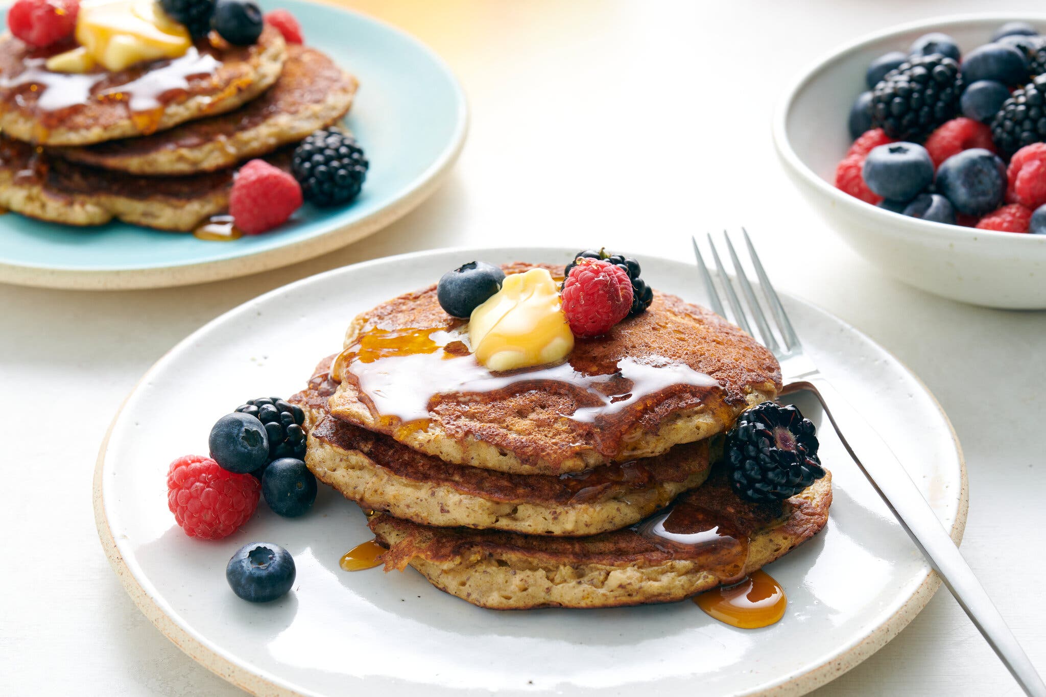 A short stack of pancakes topped with butter, berries and syrup is photographed from the side. Another serving sits just behind, next to a bowl of berries.