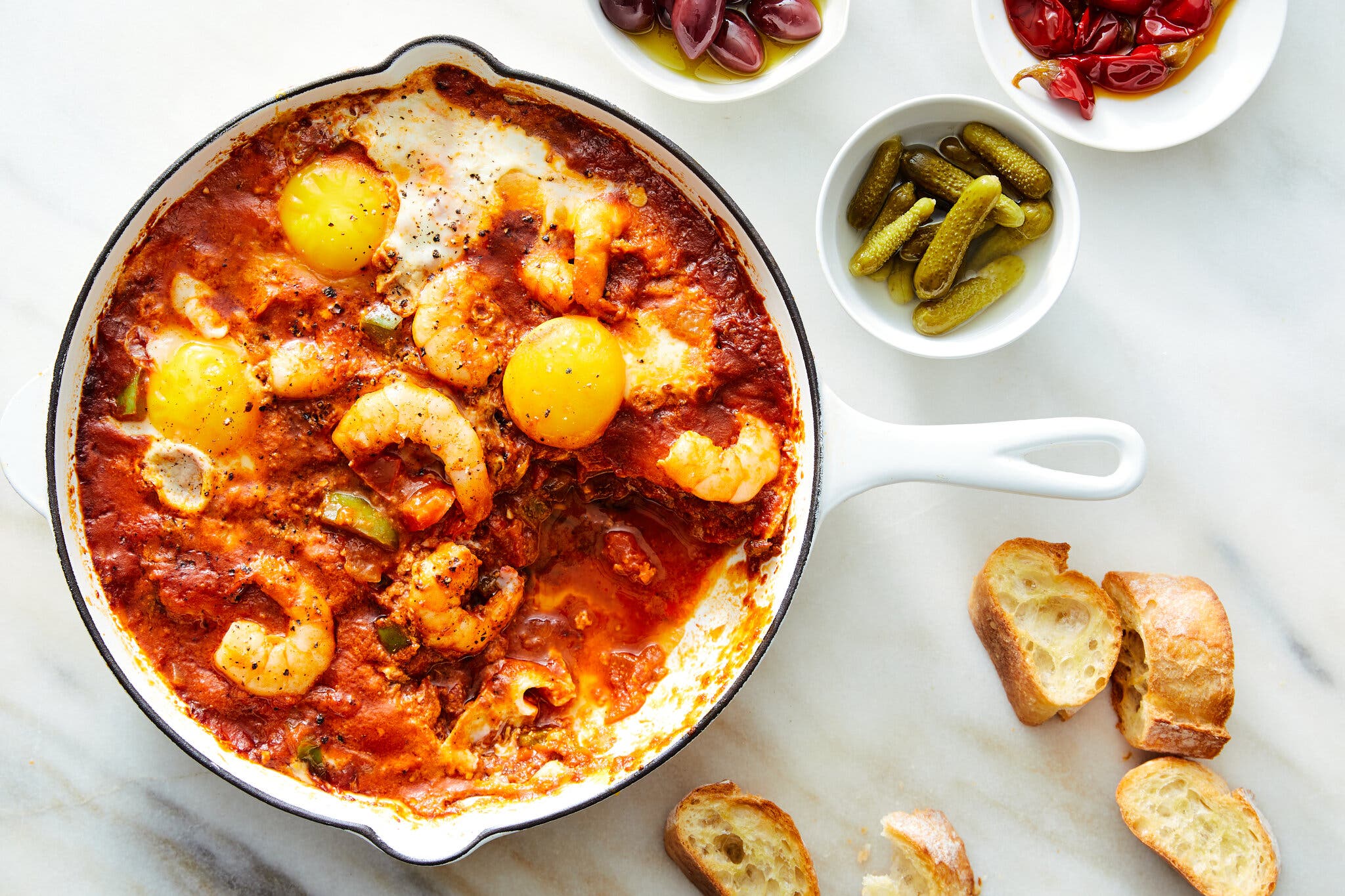 A skillet of shrimp and eggs in a chile-based shakshuka sauce, with slices of bread, cornichons, and chiles in oil alongside.