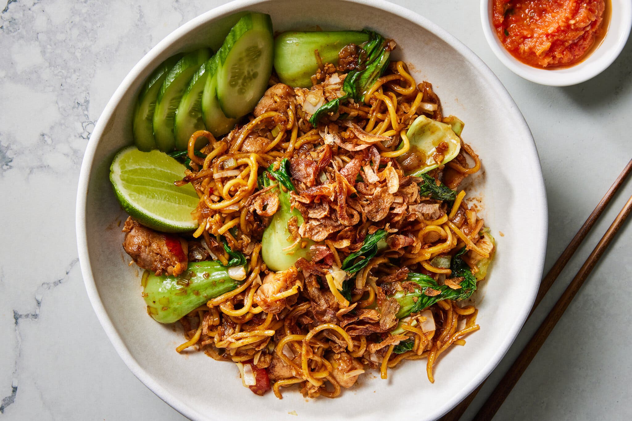 A wide, shallow bowl filled Mie Goreng, an Indonesian noodle stir-fry with bok choy and cucumber, with a small bowl of sambal alongside.