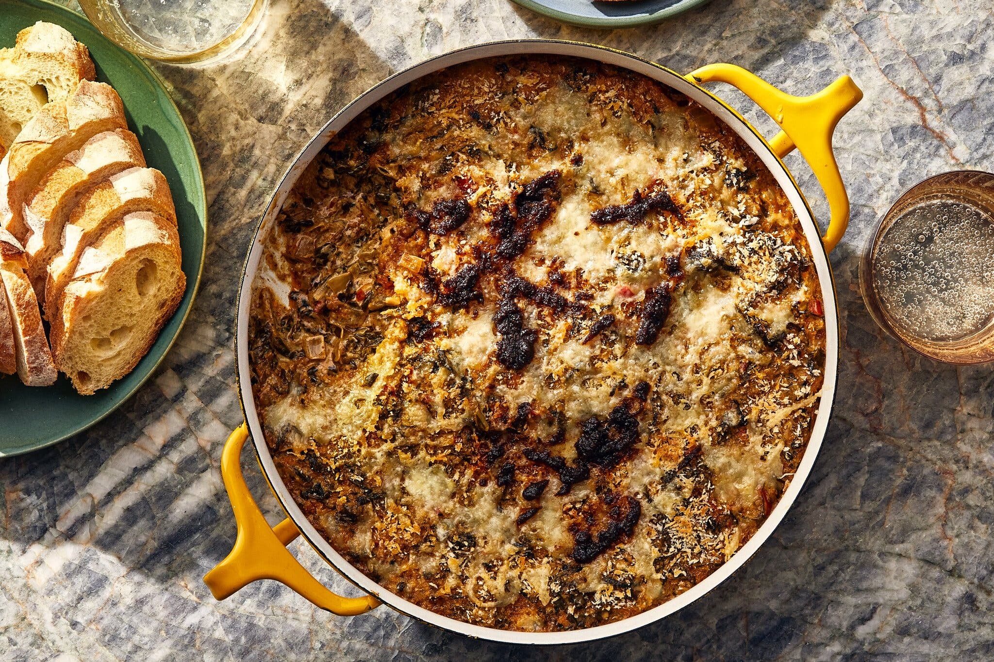 A wide skillet with a creamy collard greens dip, with sliced crusty bread and a glass of sparkling water alongside.