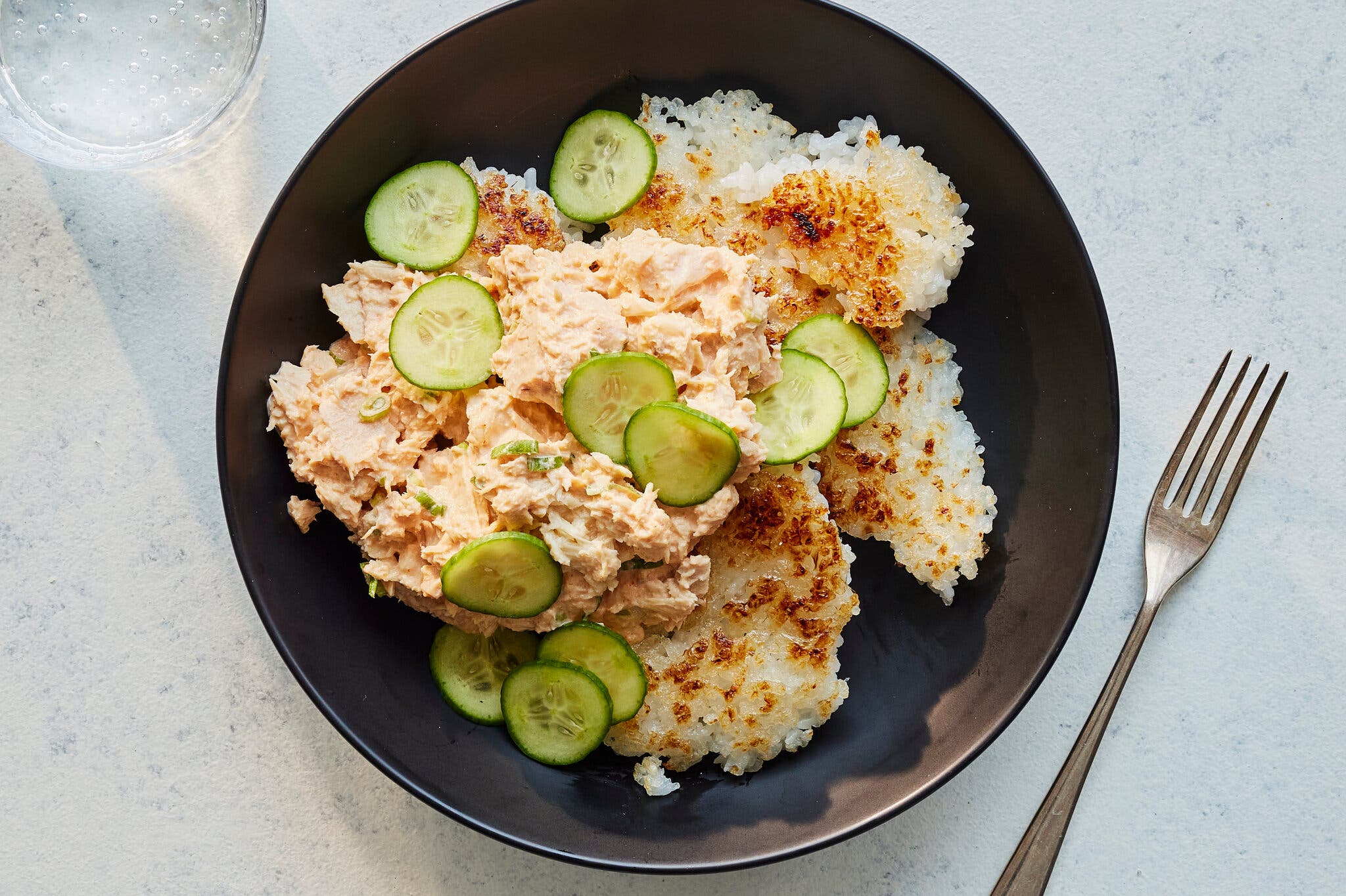 A wide, shallow bowl with crispy white rice, topped with spicy tuna salad and sliced cucumbers.