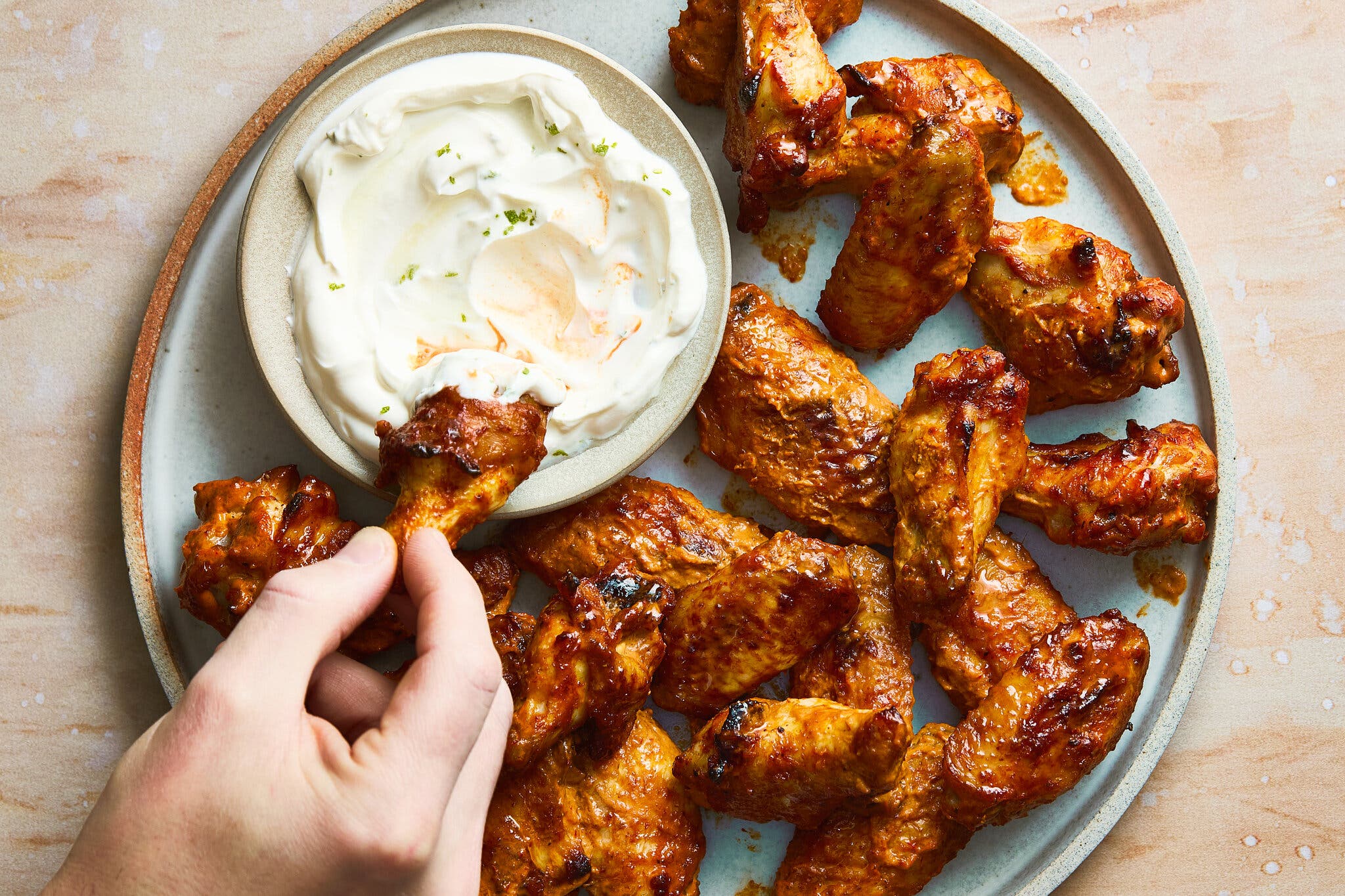 A serving platter of golden brown, saucy chicken wings, with a hand dipping one of the wings into a creamy white sauce.