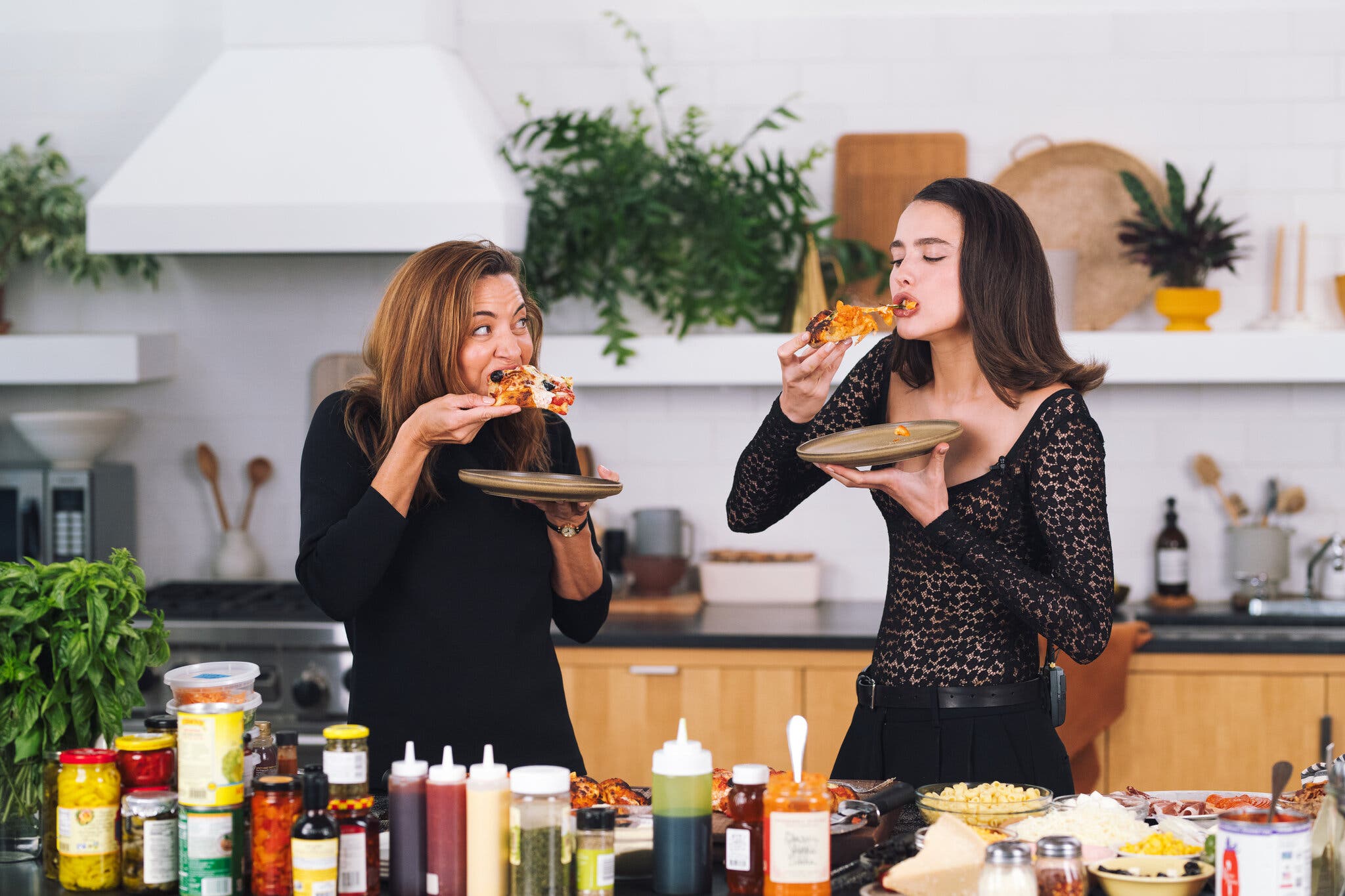Aubrey Plaza and Margaret Qualley bite into slices of pizza in the New York Times studio kitchen.