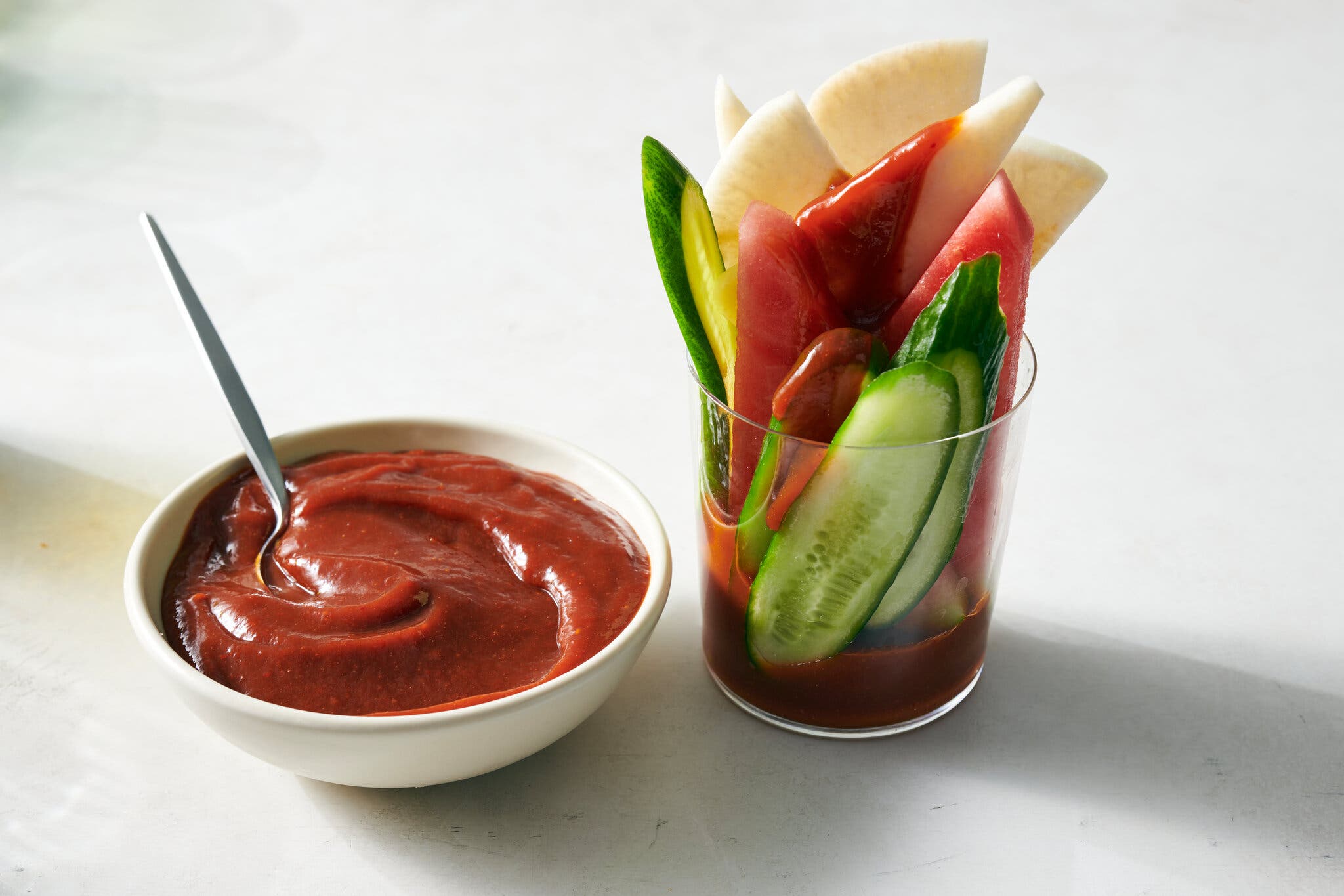 A white bowl of red chamoy sauce is next to a glass of sliced cucumber, watermelon and jicama with chamoy sauce at the bottom.
