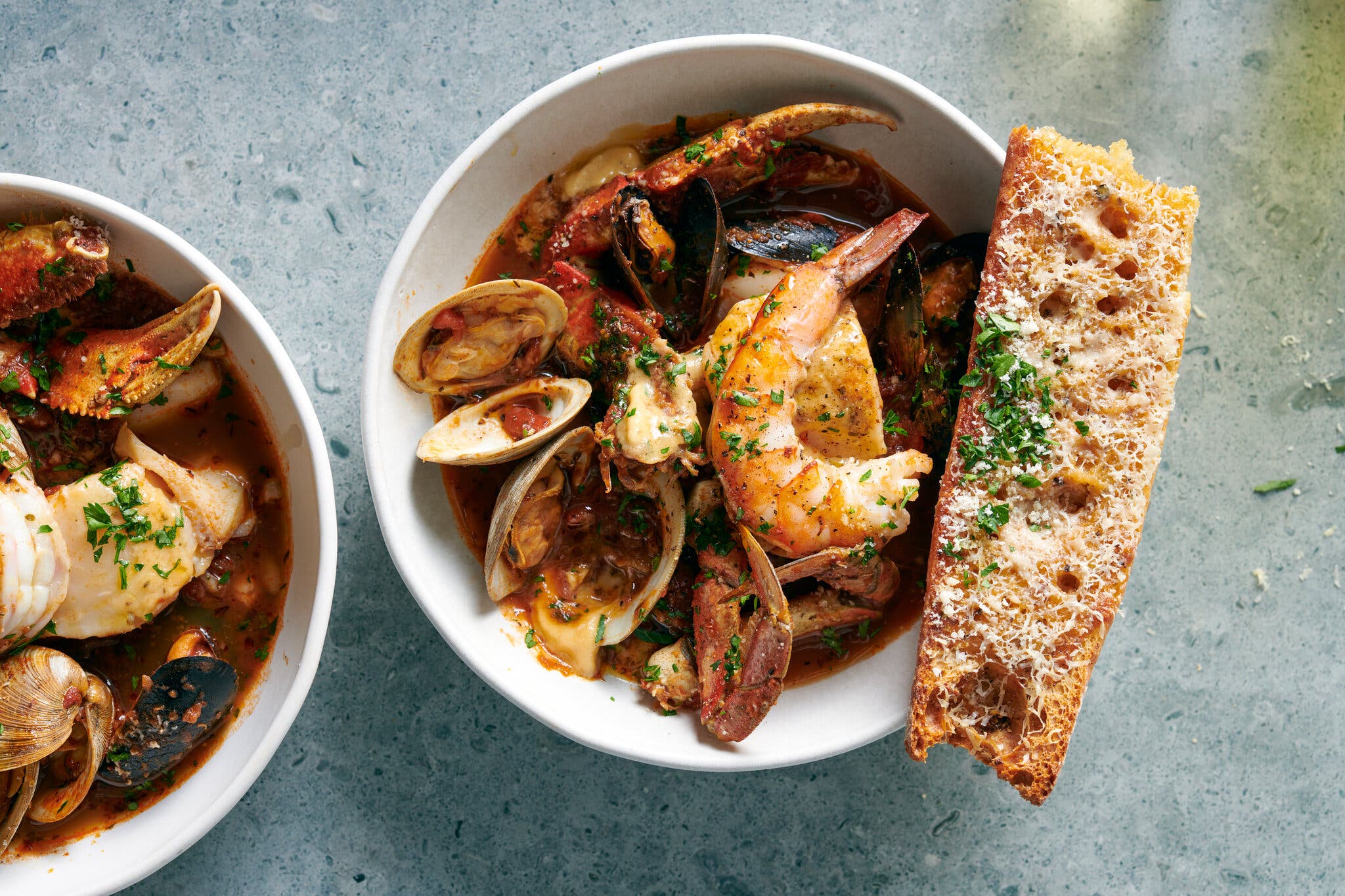 An overhead shot of a large bowl of seafood stew with crab, shrimp, clams and mussels in a reddish-brown broth, with a piece of toasted bread with cheese and herbs.