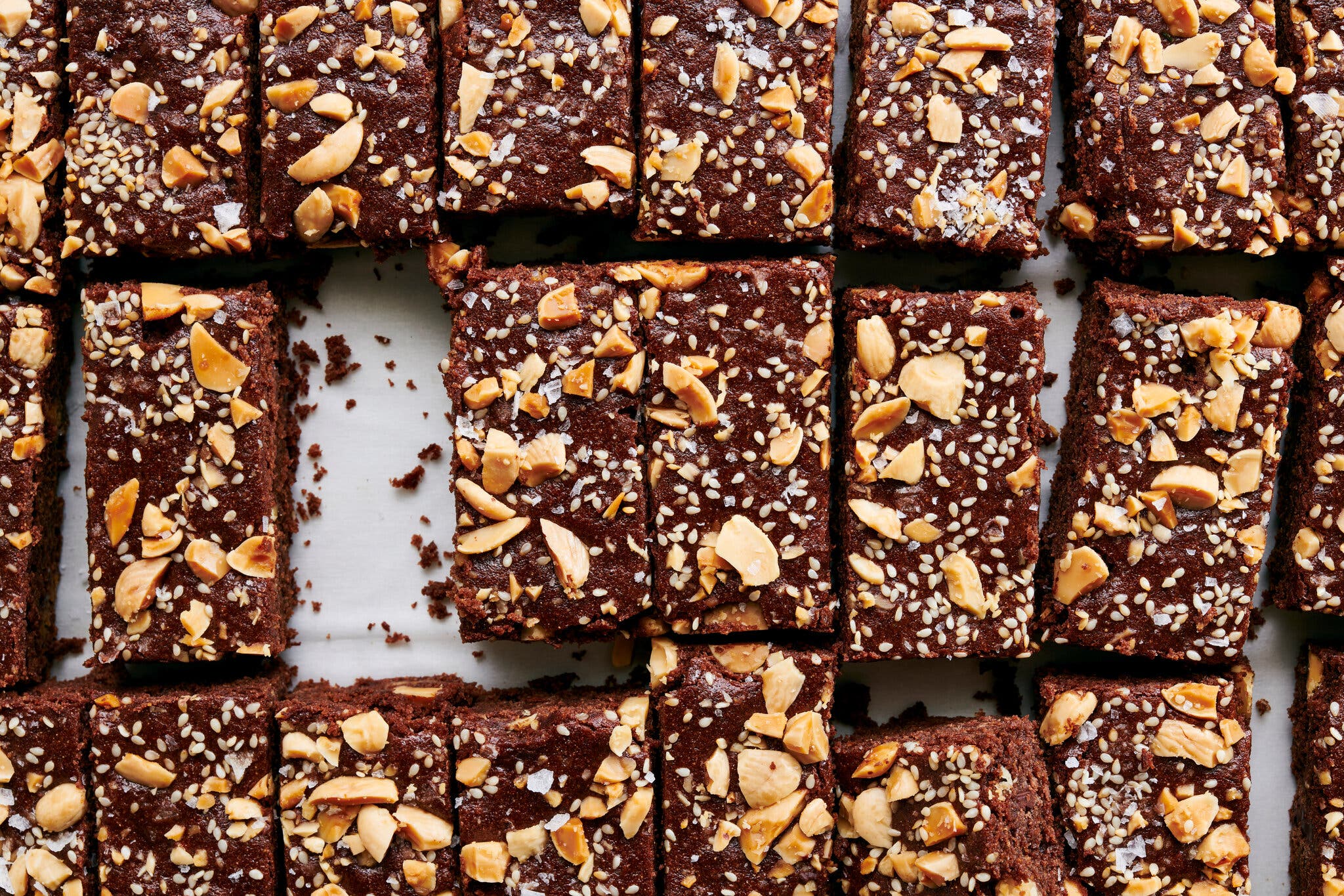 An overhead image of cut brownies topped with chopped almonds and sesame seeds on parchment paper.