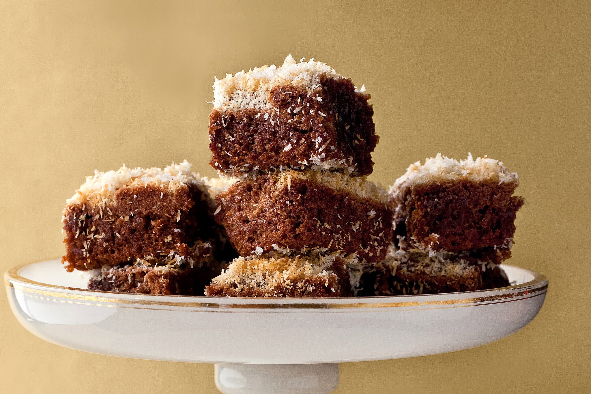 A cake plate topped with olive oil and coconut brownies against a beige backdrop.