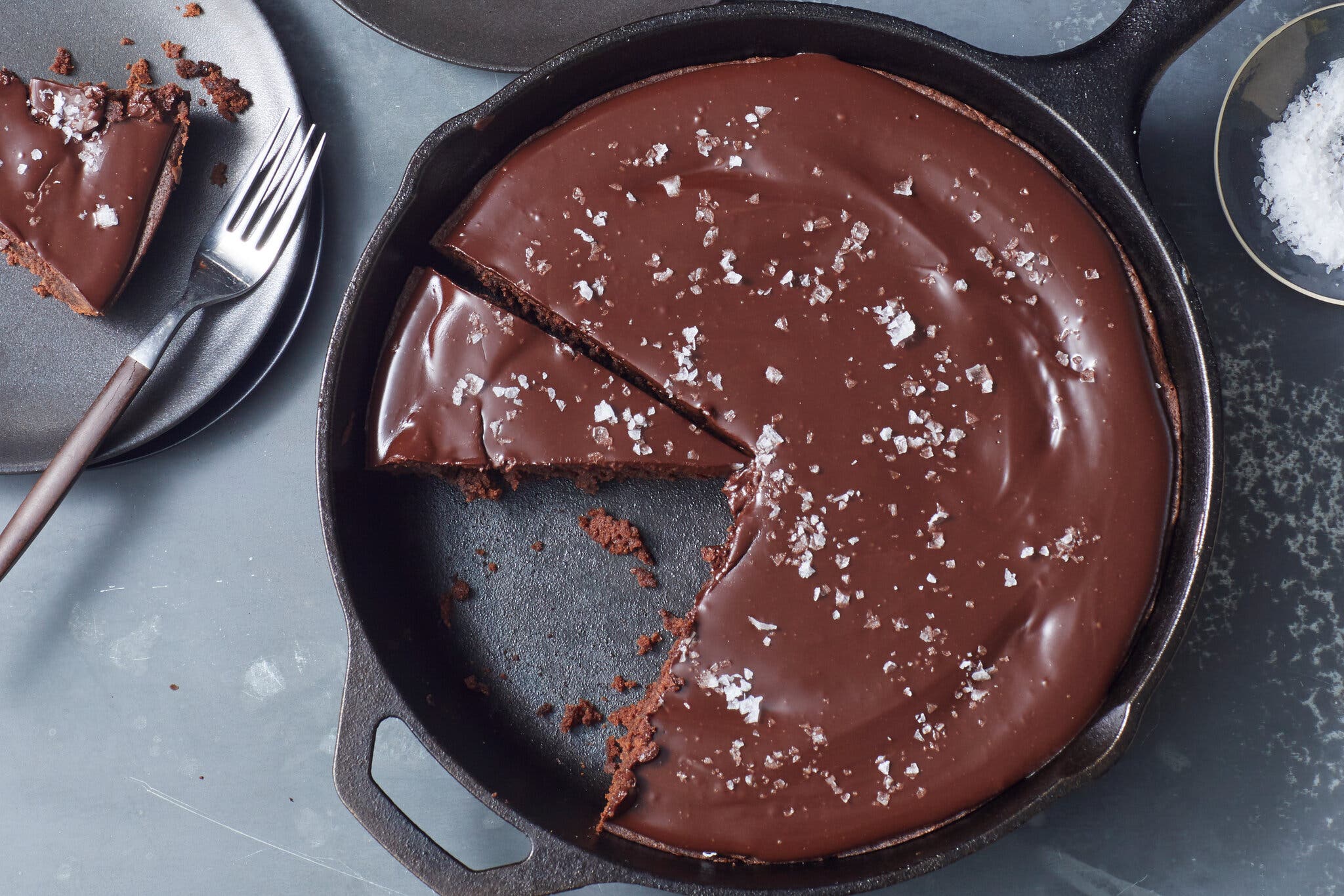 An overhead image of a cast iron skillet brownie with chocolate ganache frosting, topped with flaky salt. In the top left corner of the frame sits a plate with a slice of brownie and a fork.