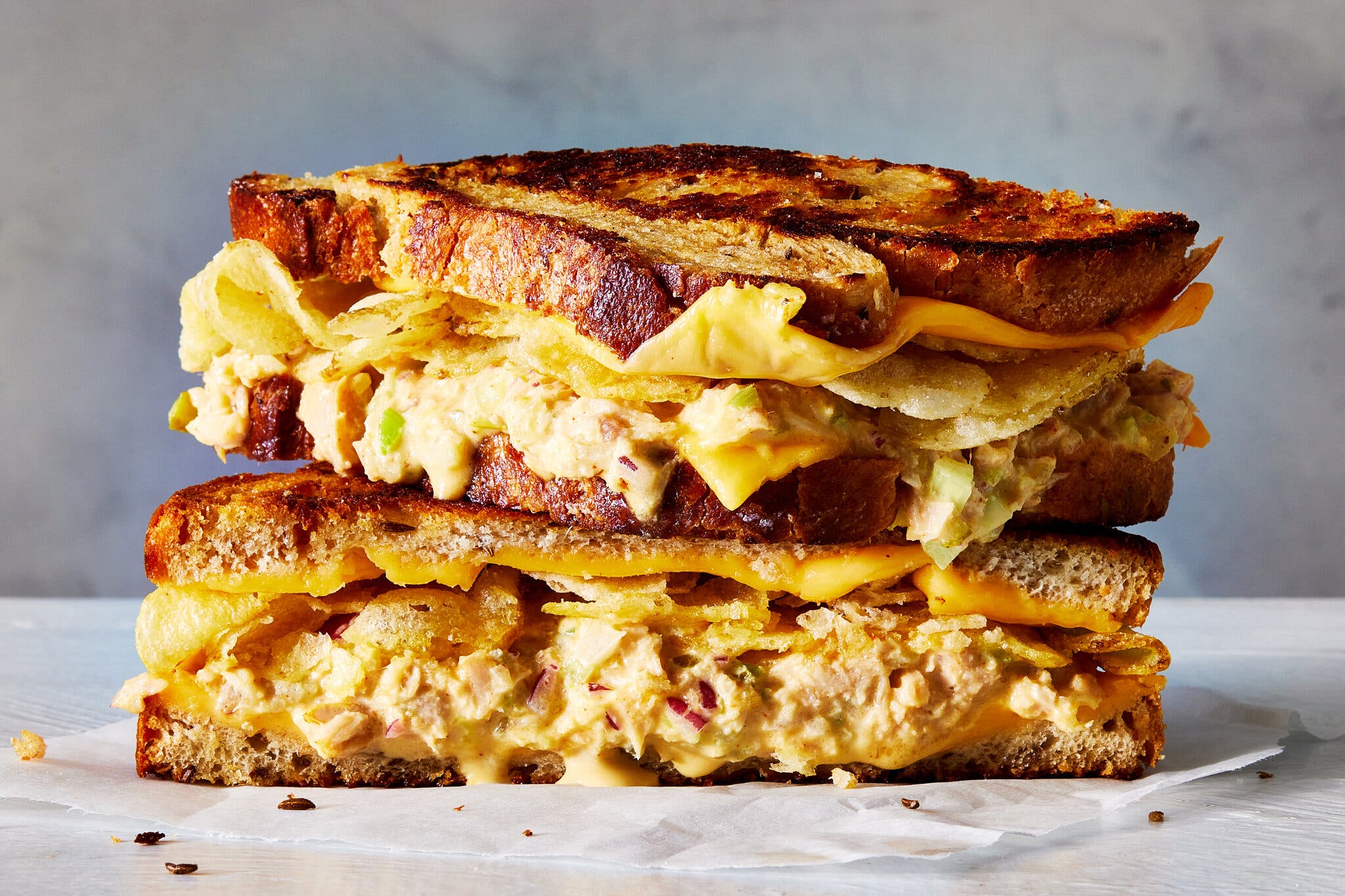 A very tall tuna melt sandwich, cut in half and stacked, with melted cheese and potato chips spilling from the sides.