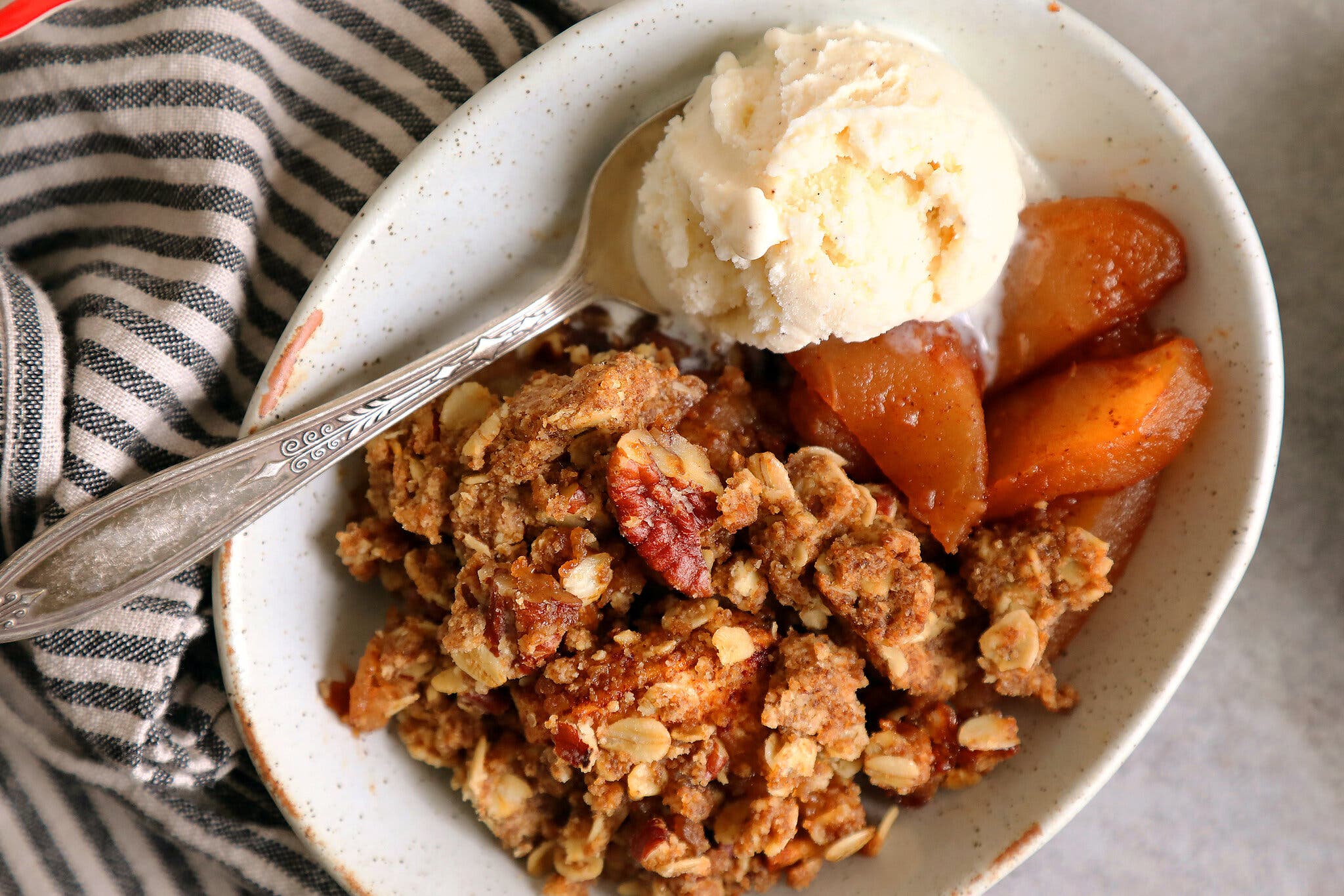 An overhead image of an oval bowl filled with apple crisp and a scoop of vanilla ice cream. A silver spoon is dipped into the ice cream. 