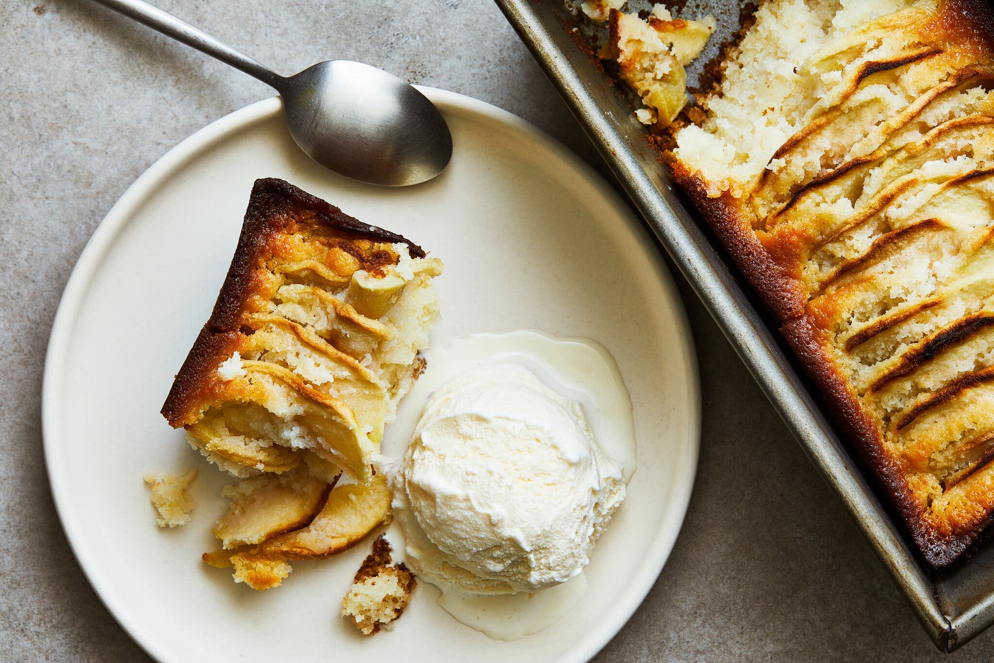 An overhead image of a baking dish filled with apple baked into a cake. A single portion and a scoop of ice cream sit next to the pan.