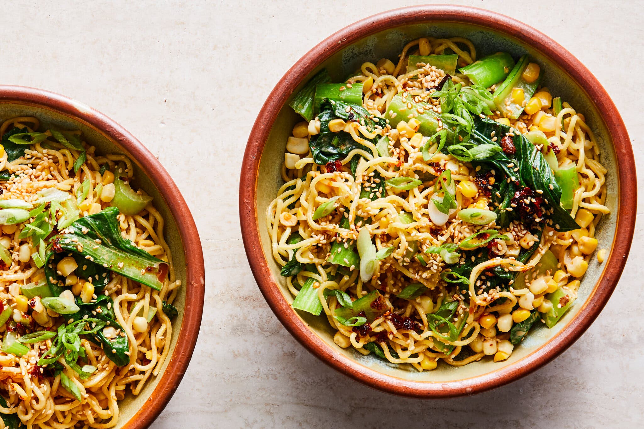 Two bowls are filled with noodles topped with green vegetables, chile and scallions.