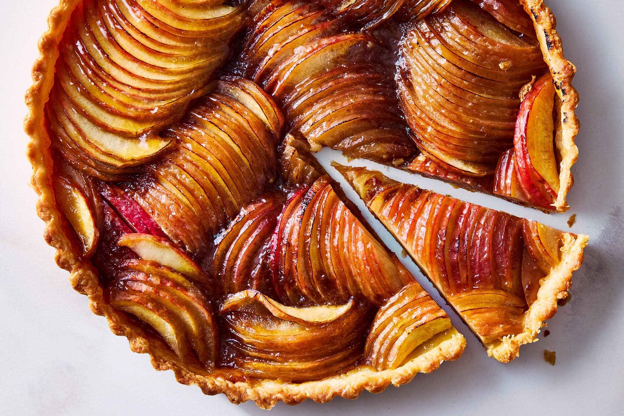 An overhead image of a tart with glossy, thinly sliced apples. The first slice is cut. 