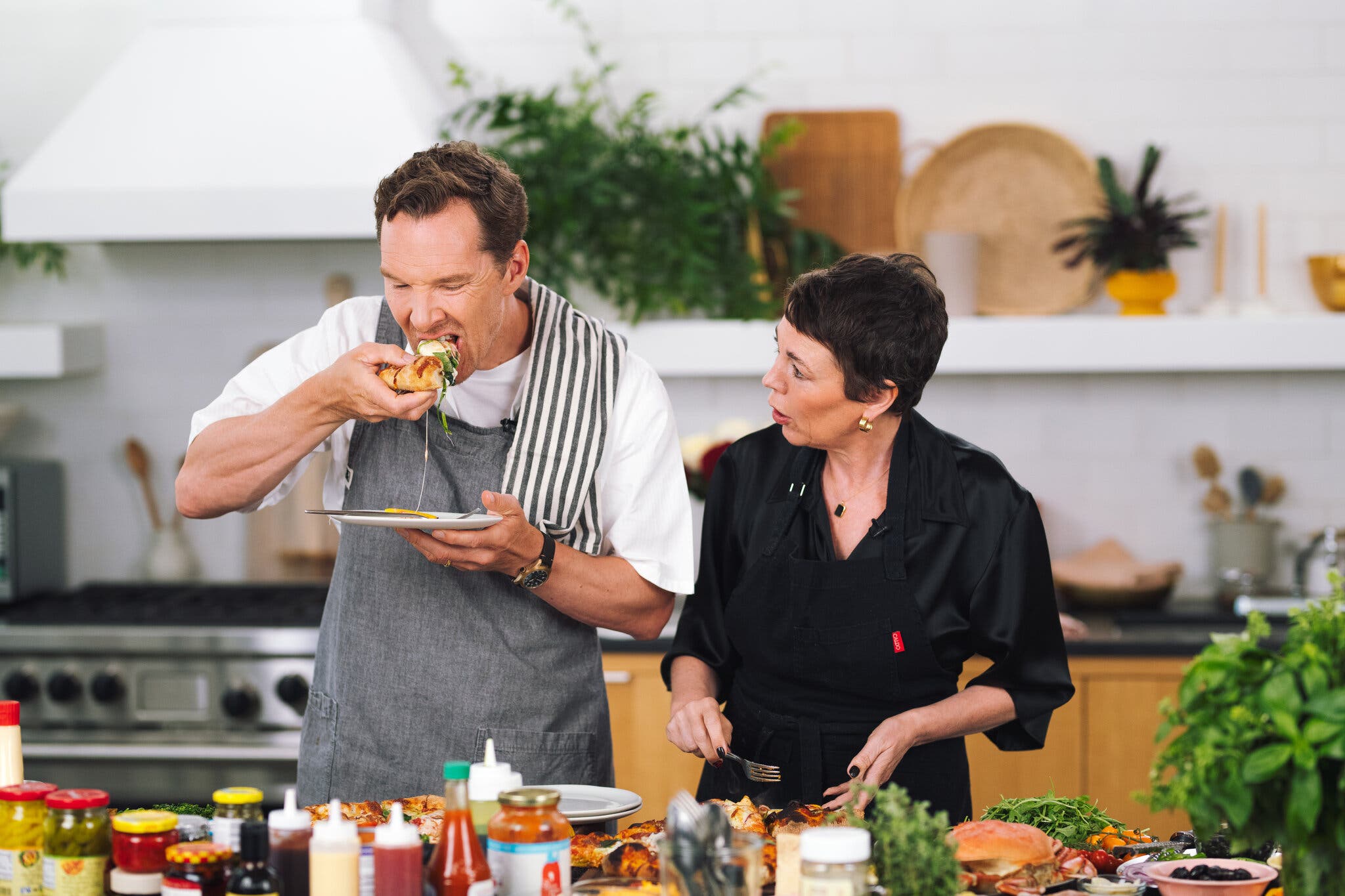 A side image of two people at a kitchen counter. A man in an apron to the left eats a slice of pizza.