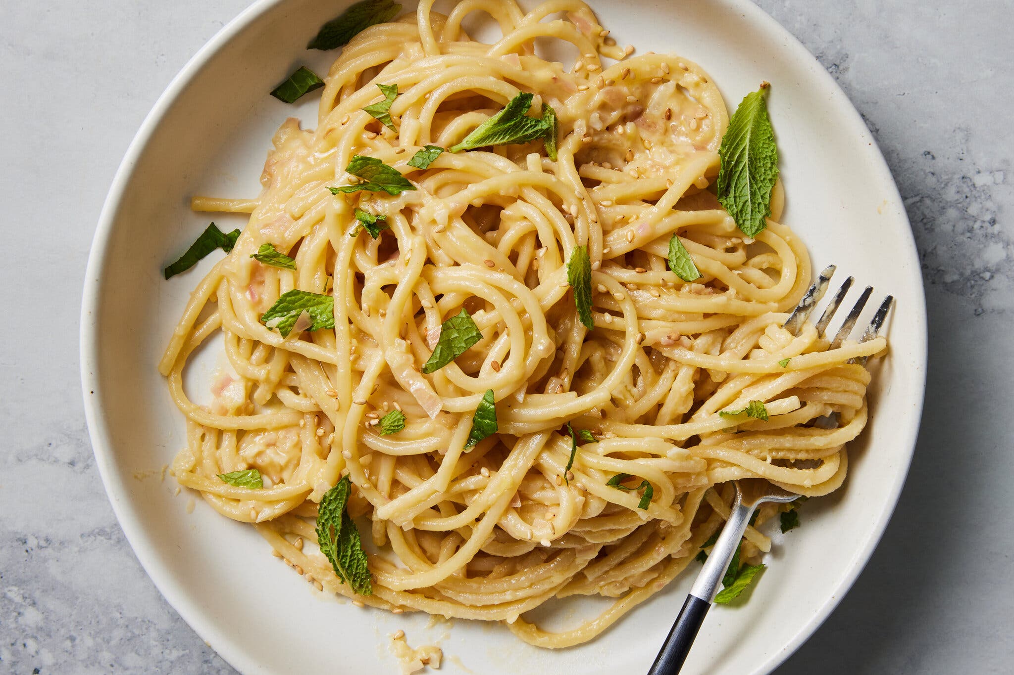 Lemony hummus pasta is shown sprinkled with sesame seeds and mint leaves on a plate with a fork.