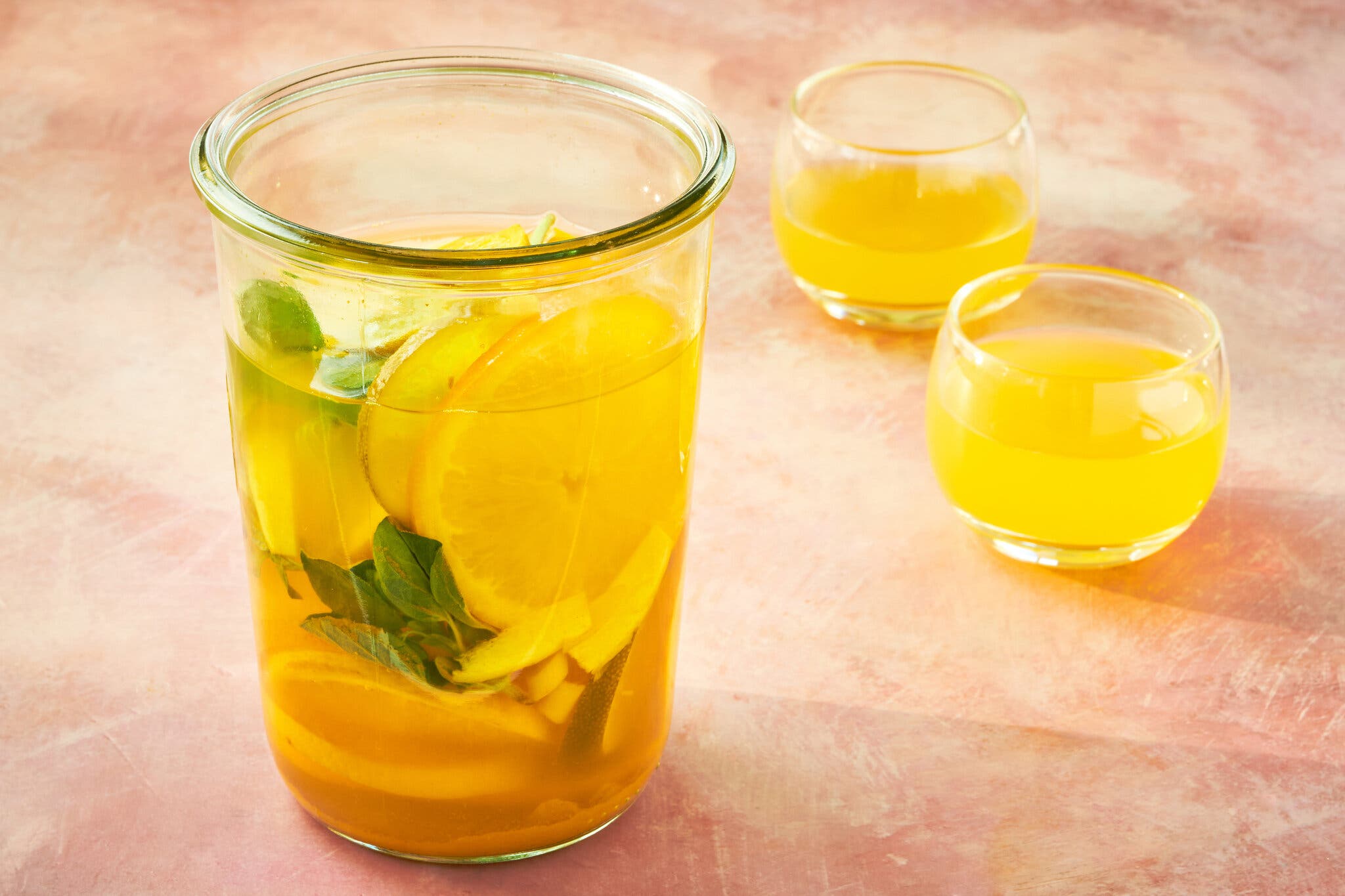 A glass container of tonic, with citrus slices and mint. There are two glasses of the orange drink nearby,