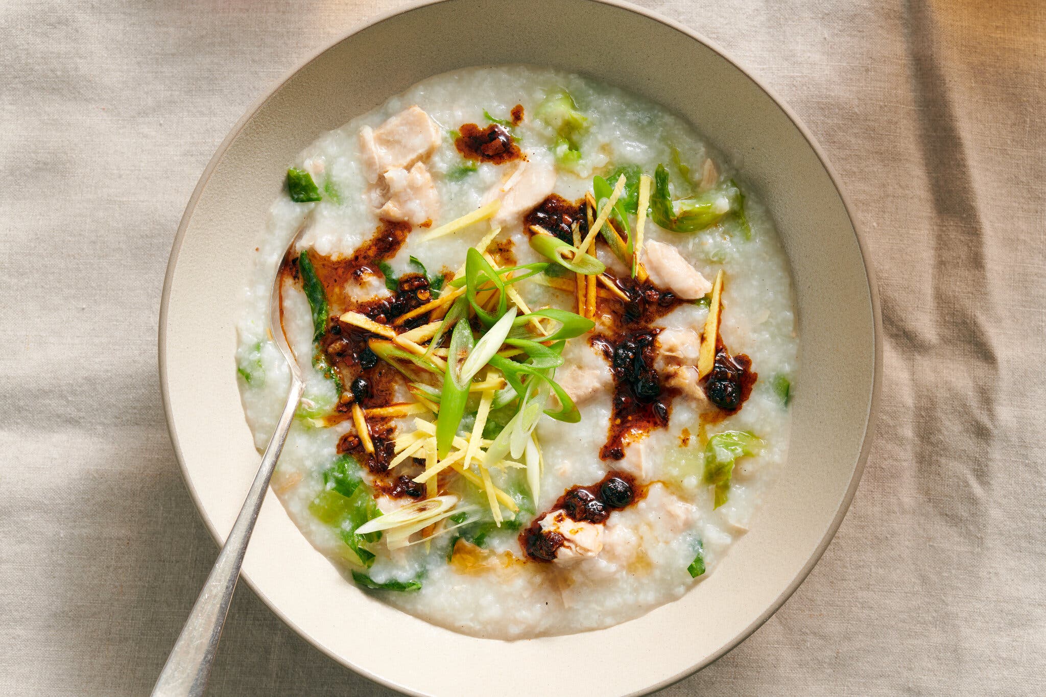 An overhead image of a bowl of congee with scallions, ginger and pieces of chicken, It’s garnished with chile oil, and there is a spoon in the bowl.