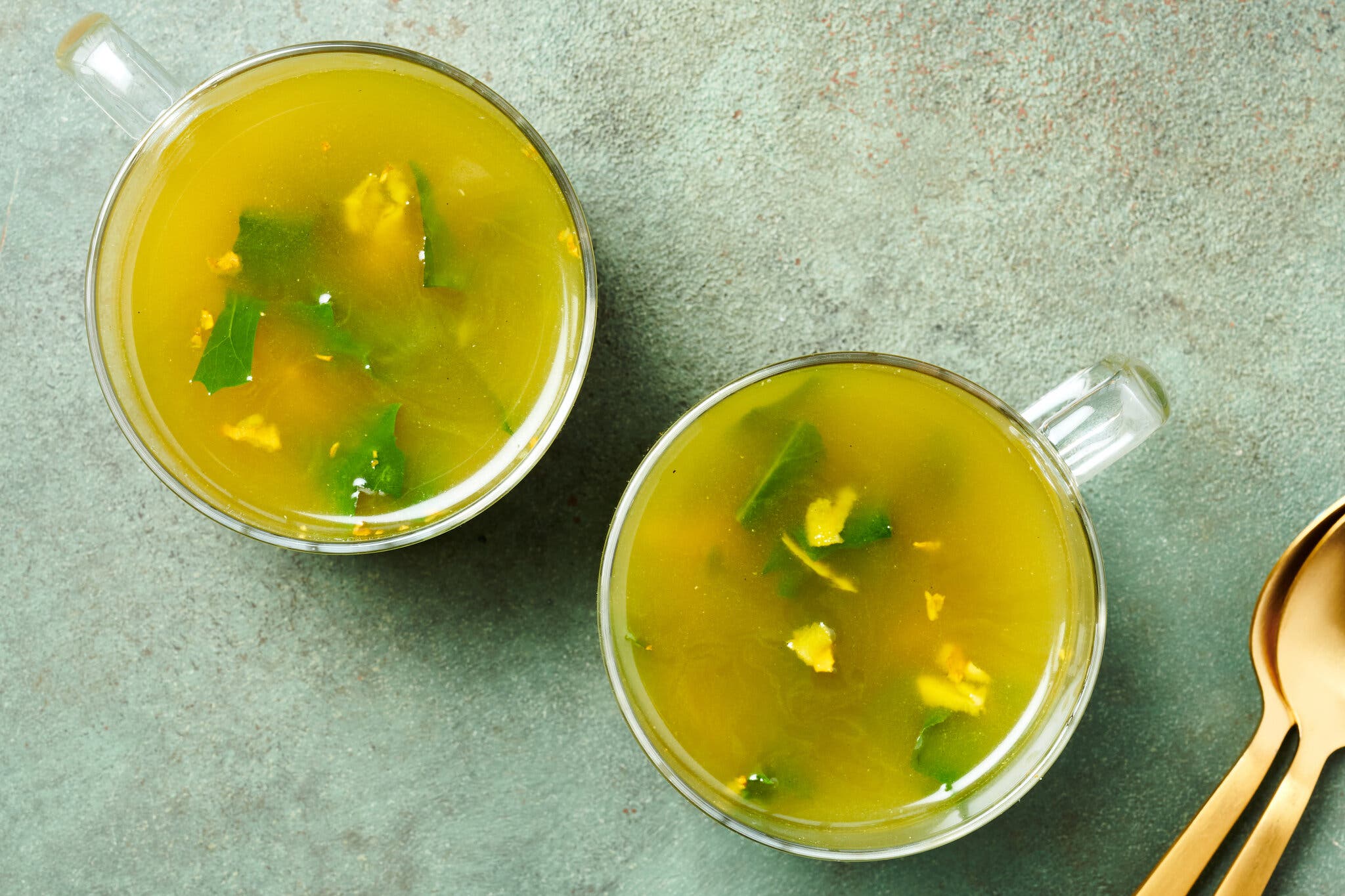 An overhead image of two glass mugs of soup, containing greens and pieces of ginger. There are two spoons nearby.