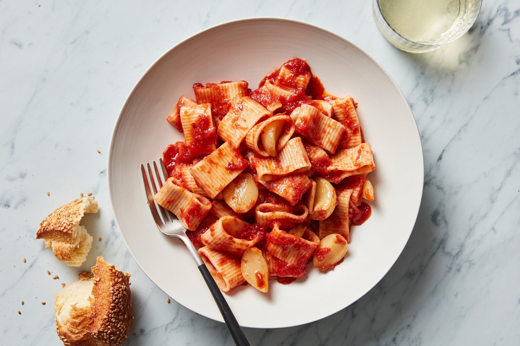 An overhead shot of a bowl of mezze rigatoni pasta with tomato sauce. There is a broken-up baguette in the bottom left corner and a fork in the bowl.