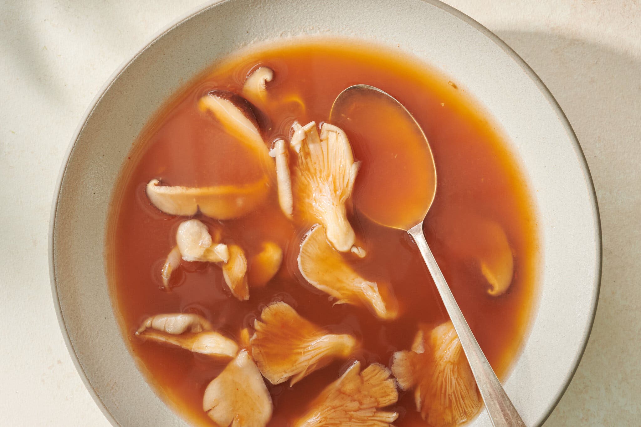 An overhead image of a bowl of clear, red soup with mushrooms in it. There is a spoon in the bowl.