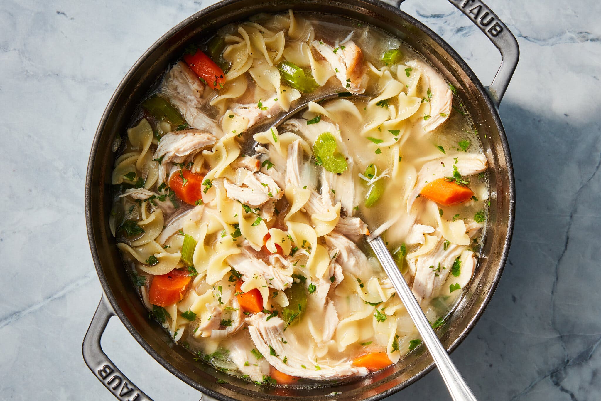 An overhead image of a Dutch oven full of chicken noodle soup, with egg noodles, shredded chicken, celery and carrots. There is a metal ladle in the pot.