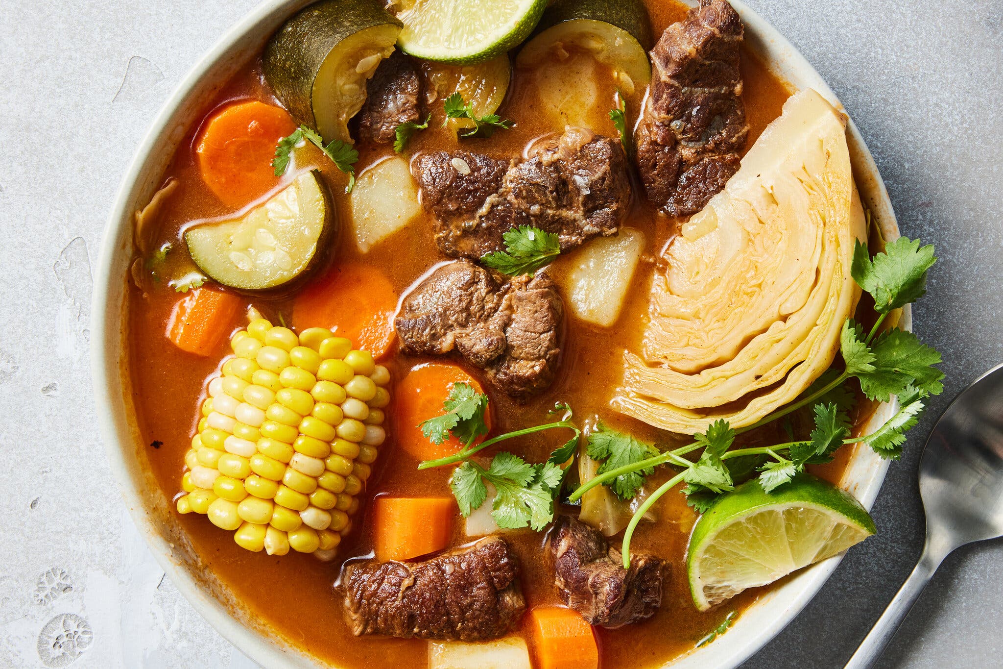 An overhead image of a bowl of soup with beef shank, carrots, zucchini, cabbage and corn, garnished with cilantro and a lime wedge.