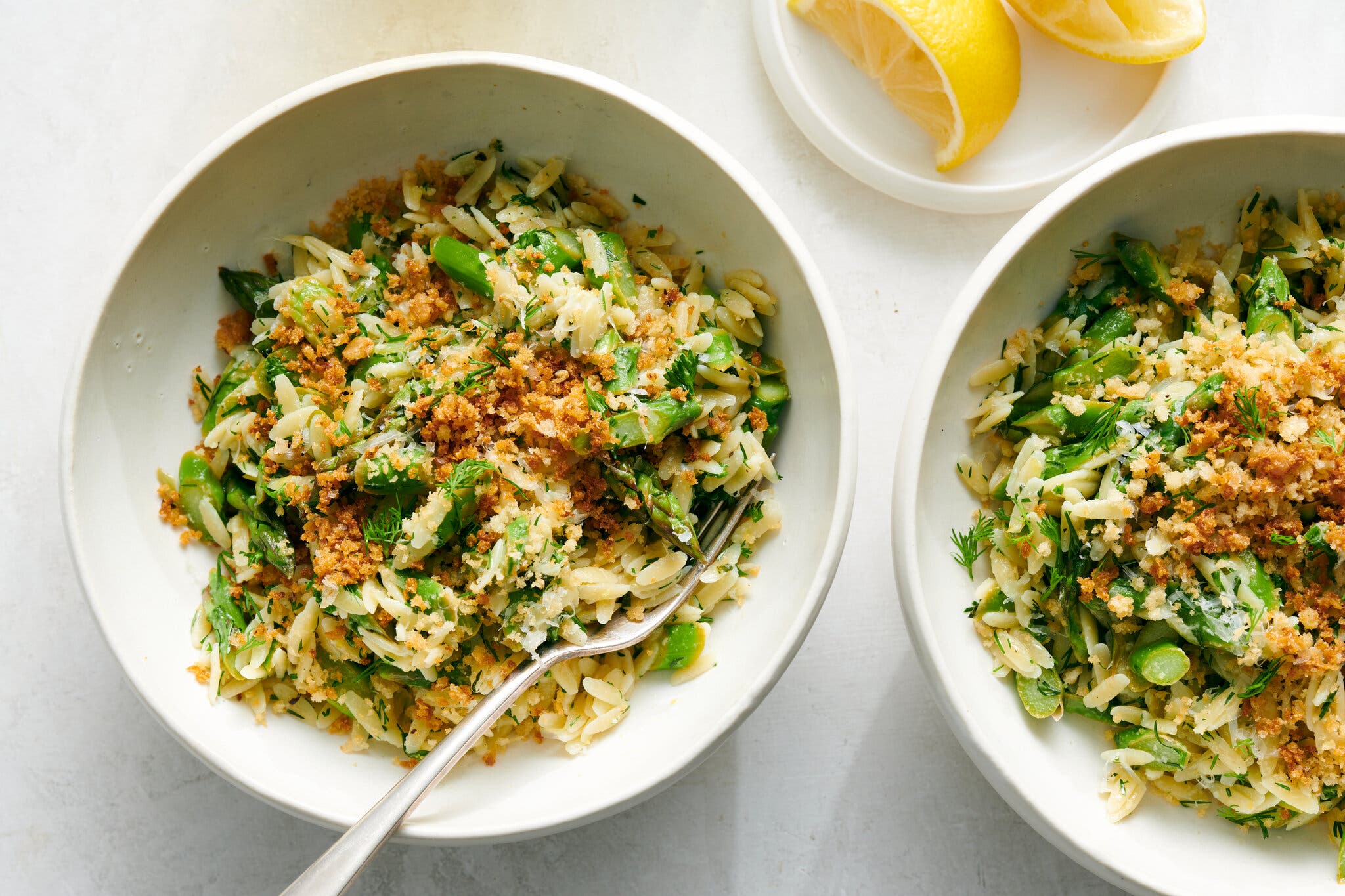An overhead image of two bowls of orzo with asparagus and bread crumbs. There is a small dish with lemon wedges towards the top, and a fork in the first bowl.
