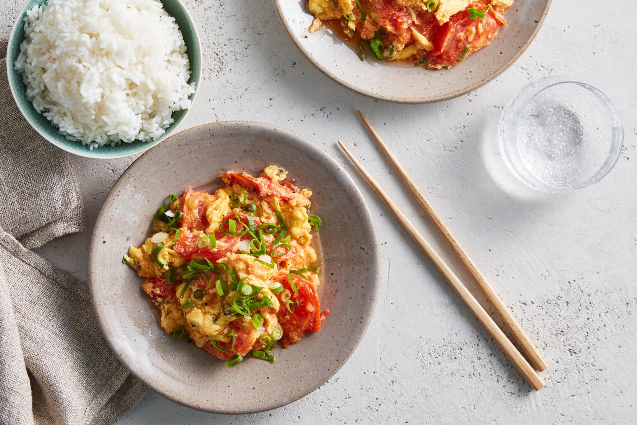 An overhead shot of a bowl of Chinese stir-fried tomatoes and eggs, garnished with sliced scallions. There is a bowl of rice, a pair of chopsticks and a glass of water nearby.