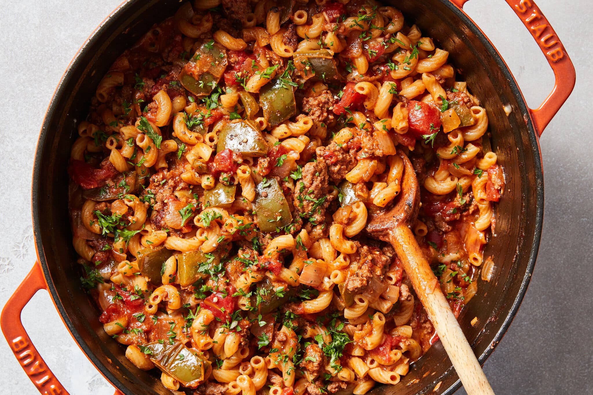 An overhead shot of a Dutch oven full of goulash, with elbow macaroni noodles, green and red bell peppers and ground beef. There is a wooden spoon in the pot.
