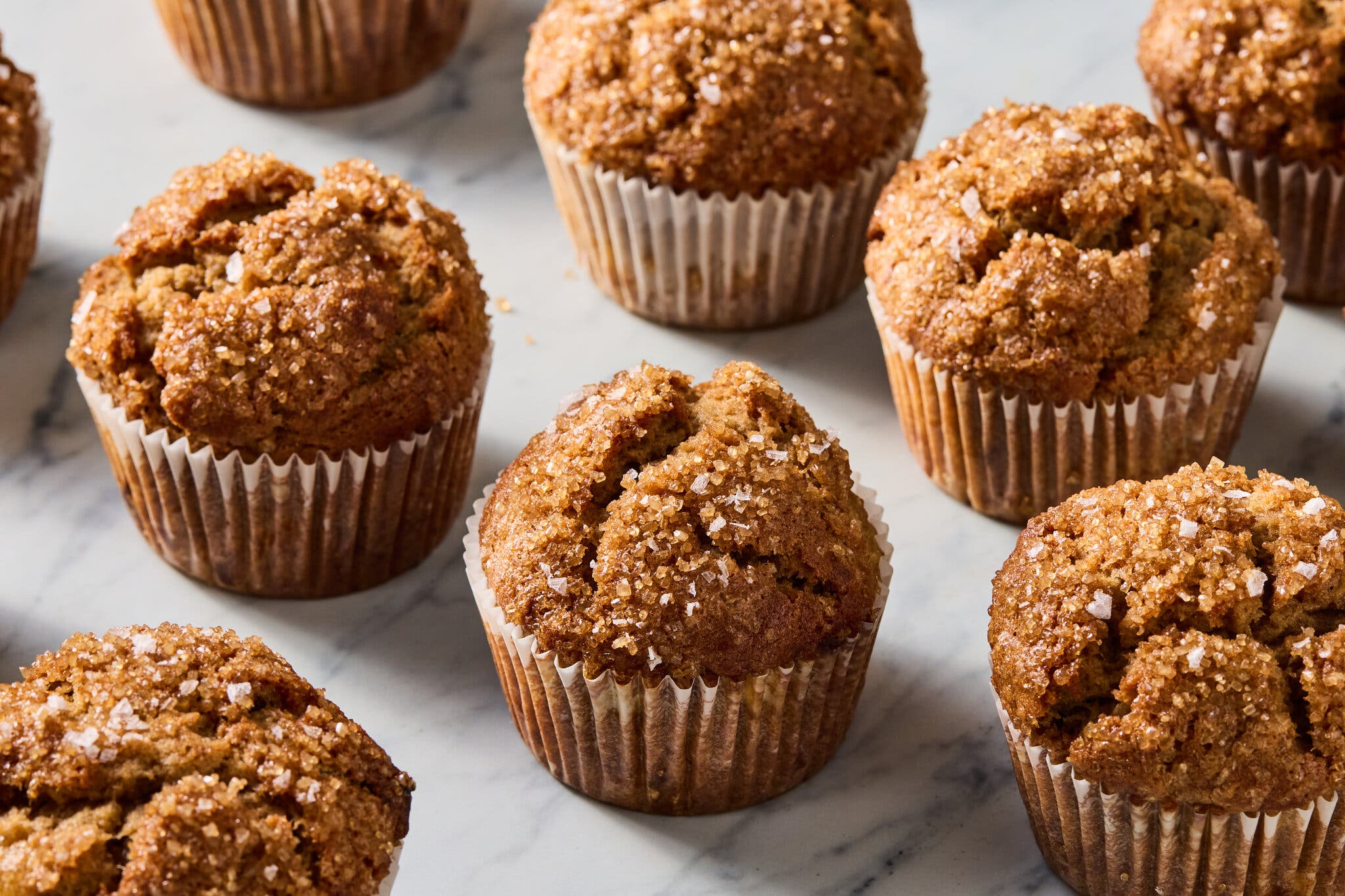 Banana muffins, garnished with turbinado sugar and flaky salt, wrapped in muffin liners, sit on a marble countertop.