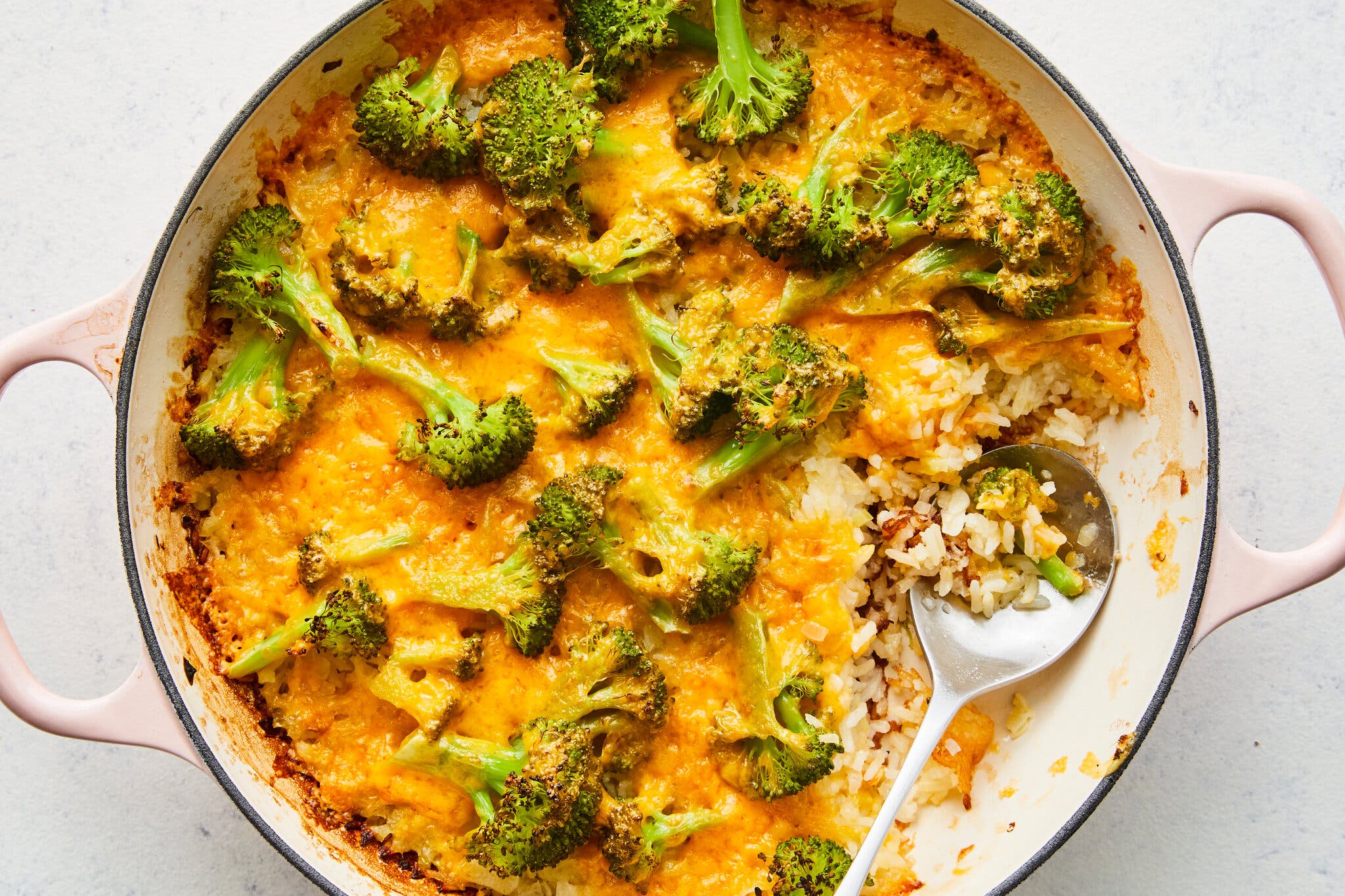 An overhead shot of a Dutch oven of rice, covered in crackly cheese and pieces of broccoli. There is a spoon in the bowl.